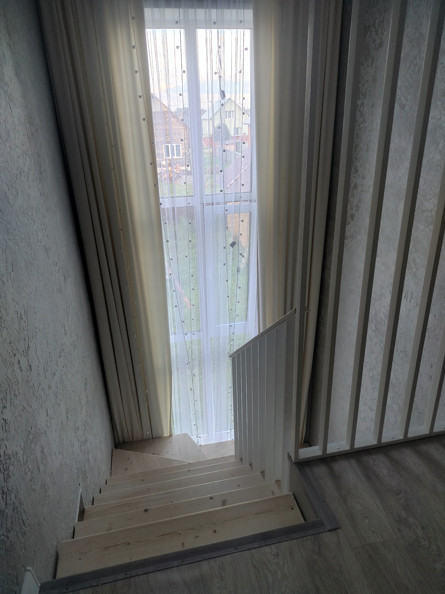 The image shows a staircase in an interior, likely a residential building in Pomryaskino, Russia. The staircase is made of unfinished wood and ascends towards a large window covered by sheer curtains with decorative lights. To the left of the stairs, the wall has a textured gray finish. To the right, there is a wall with vertical ribbed paneling. Beyond the window, houses and greenery are visible, suggesting an outdoor view. The flooring at the bottom of the stairs appears to be laminate. No people are present in the image.
