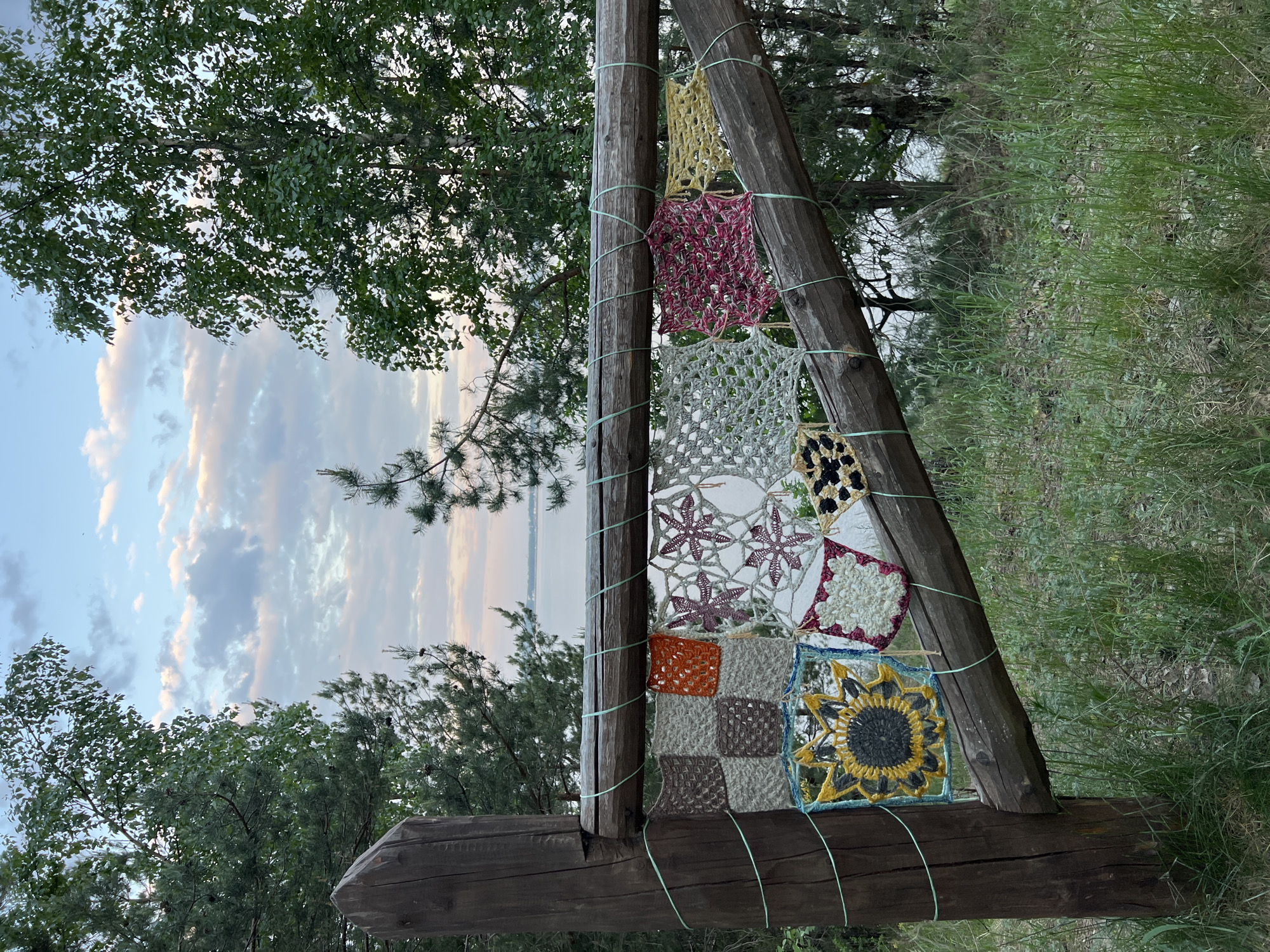 The image presents a wooden triangular structure with a collection of crocheted squares hanging from it. The structure is set against a backdrop of trees and a cloudy sky, with green grass visible to the right. The crocheted squares exhibit various patterns and colors, including floral motifs and geometric designs. The scene appears to be outdoors, possibly in a rural or natural setting. While the location is specified as Noviy, Russia, there is no evidence in the image that confirms the location.