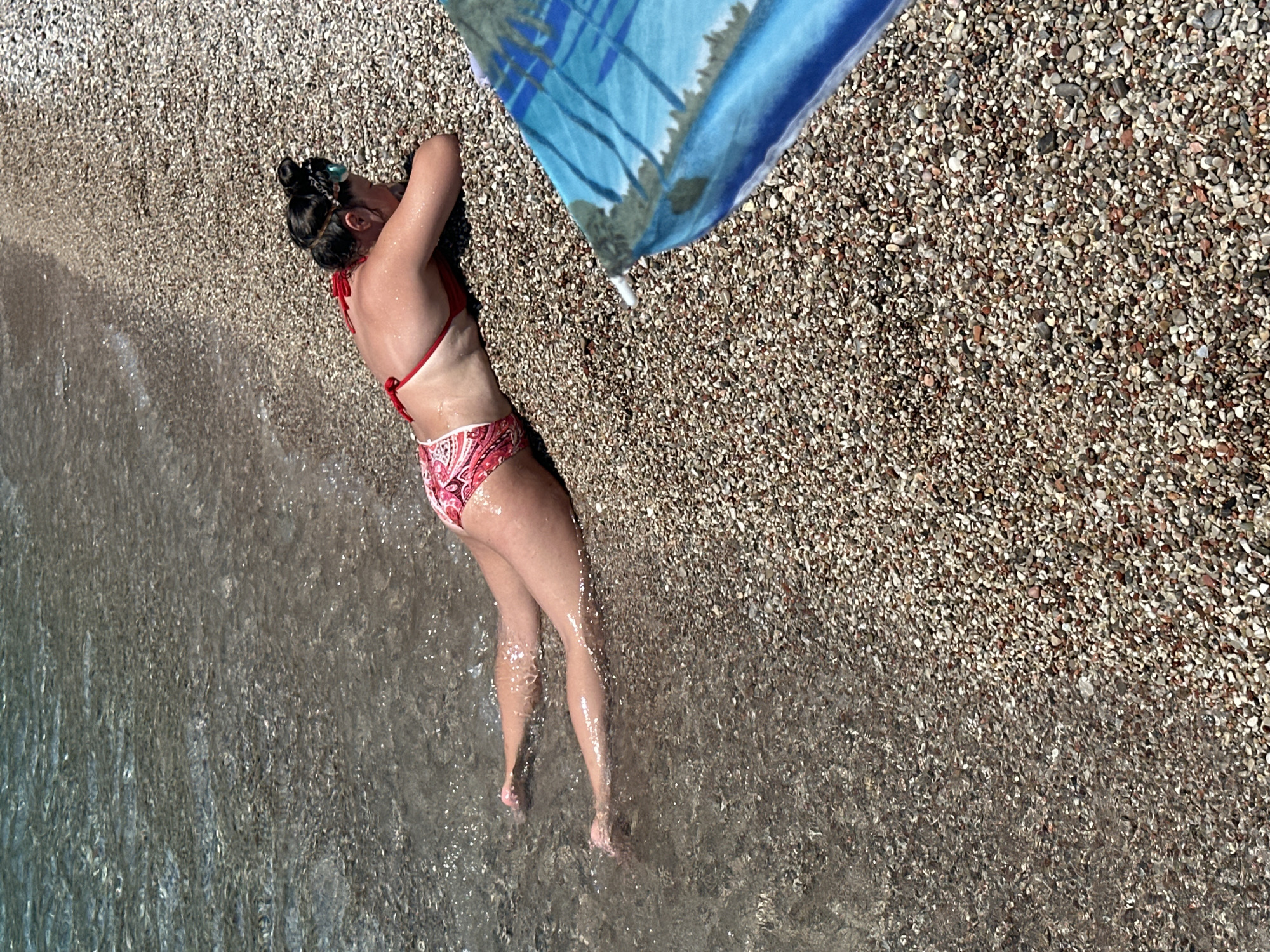 In Mirište, Montenegro, a woman with dark hair pulled into a bun lies face down on a pebble beach, her body partially submerged in the clear, shallow water. She wears a red bikini with a paisley pattern. The beach is covered in small, multicolored pebbles, and the water is calm and transparent, revealing the smooth stones beneath the surface. To the right of the frame, a blue beach umbrella with a palm tree design is partially visible, casting a shadow on the pebbles. The overall scene depicts a tranquil beach environment on a sunny day.