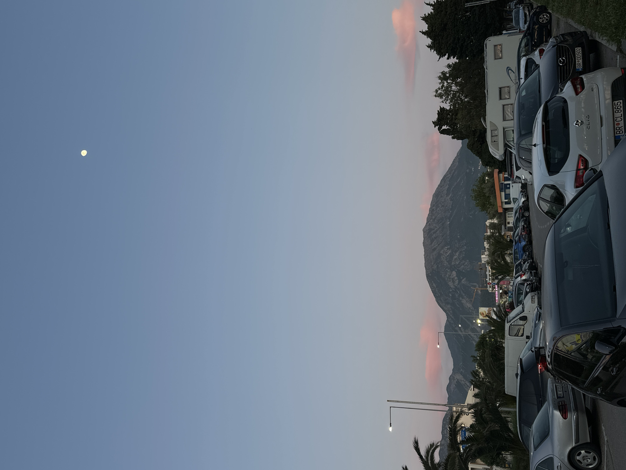 The image captures a serene dusk or dawn scene in Bar, Montenegro, with a clear blue sky transitioning into lighter tones near the horizon. Delicate pink and orange clouds are scattered, and a bright, distinct moon is visible in the upper left quadrant of the sky.
The right side of the frame is filled with a parking lot containing numerous parked cars of various models and colors, including a white camper van/RV, a white Renault Clio, and several sedans. European license plates are visible, confirming the location as Bar, Montenegro, with "BR CL 865" and "BR 00502" discernible on different vehicles.
In the mid-ground, majestic mountains rise, their rugged silhouettes set against the sky. Scattered buildings are present at the base of the mountains, and green trees, including some palm trees, are interspersed among the vehicles and structures. Several street lamps are visible, with at least one illuminated, enhancing the evening atmosphere. A faint sign in the distance appears to read "TABACO SHOP". No people are explicitly visible. The scene evokes a tranquil transition period between day and night in a coastal town.