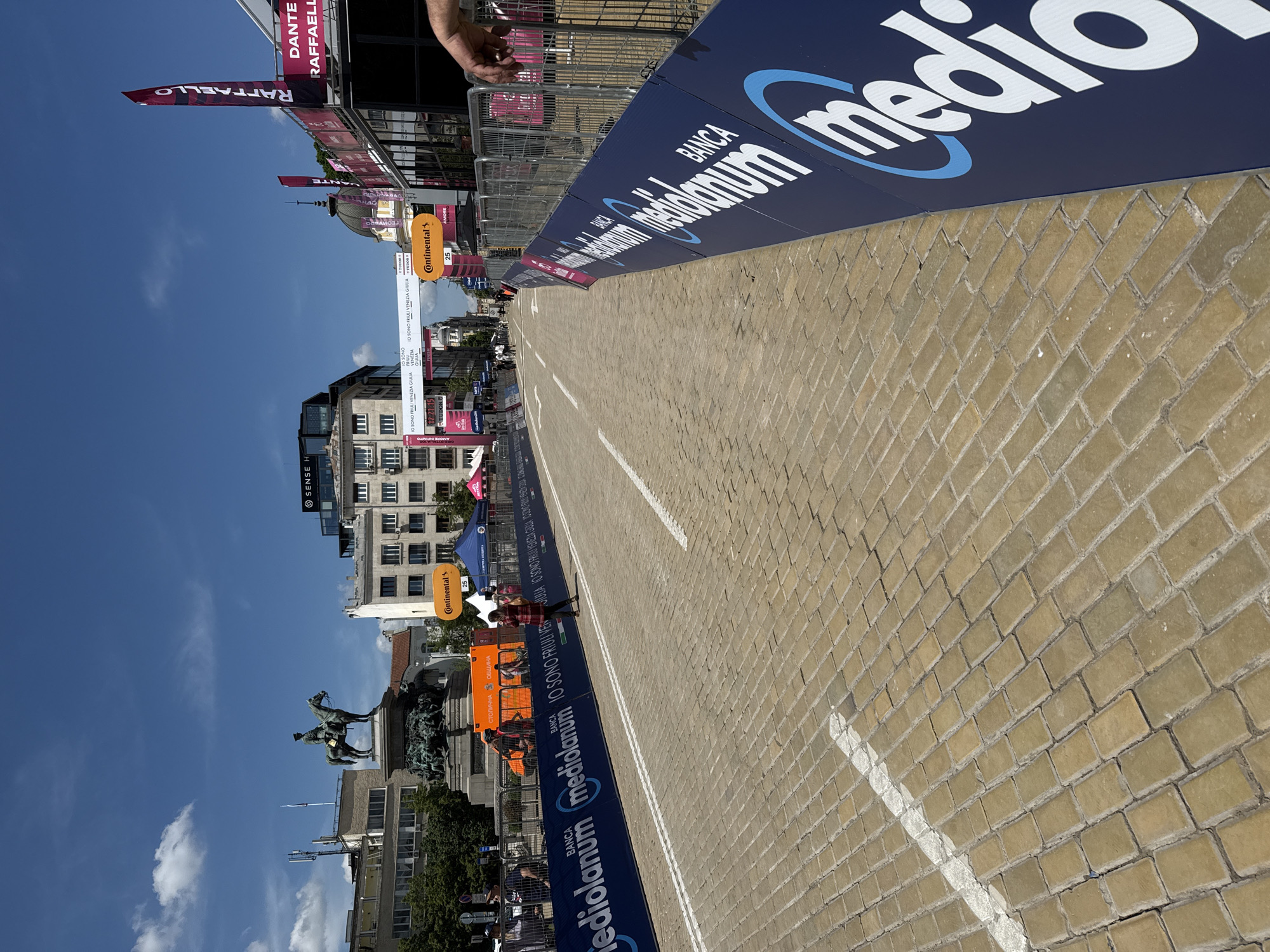 The image depicts a street in Sofia, Bulgaria, likely during a cycling race or event. The road is paved with yellow bricks and has white lane markings. Blue banners with "Banca Mediolanum" are visible on both sides of the street. Buildings and banners line the street in the background, with clear blue skies above. A statue of a horse and rider is visible on the left side. Several individuals are seen along the edges of the street. Banners and signs are affixed to buildings and fences. The time visible is 21:15.