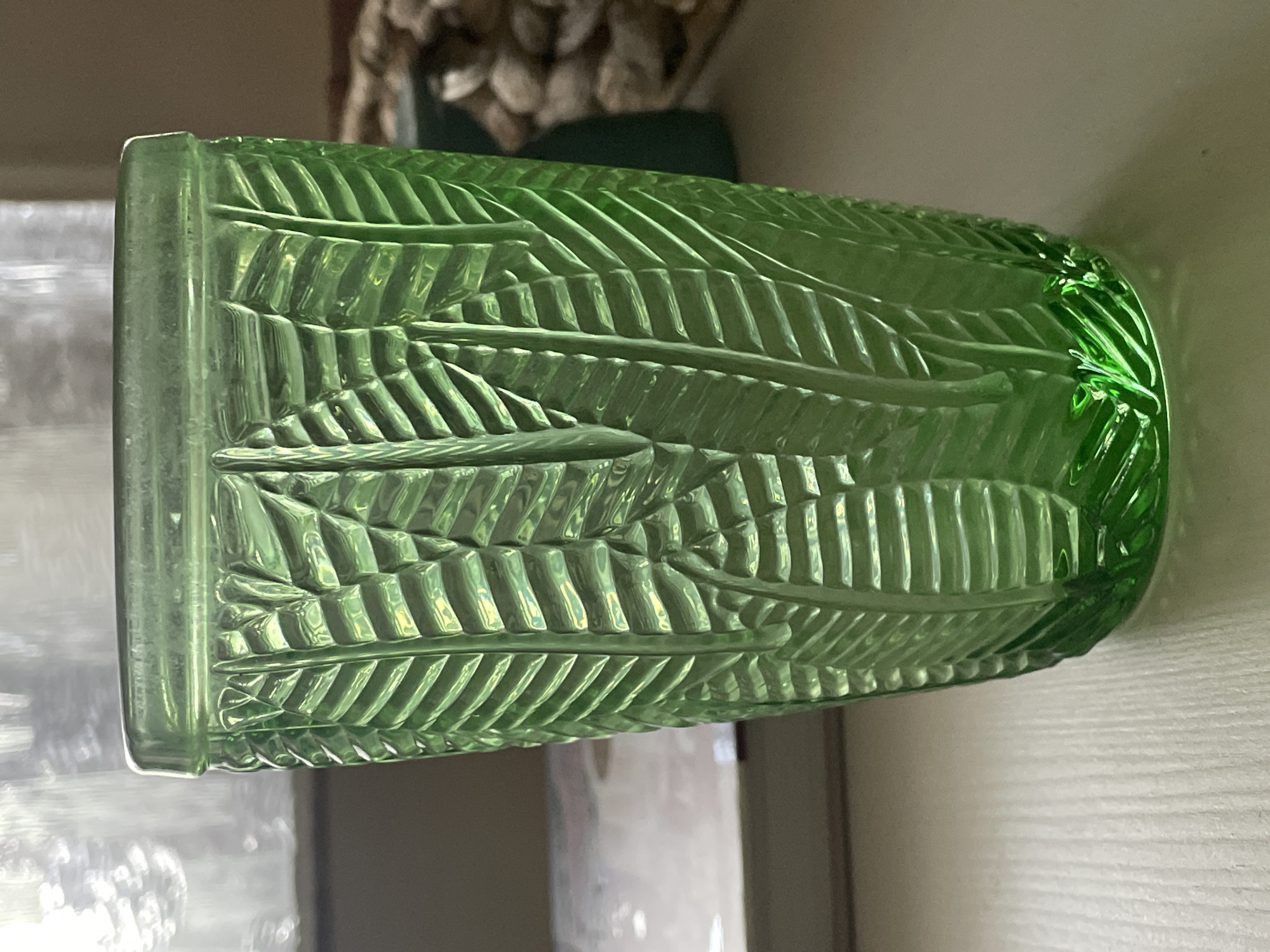 A close-up, eye-level shot shows a tall, green glass tumbler with an embossed leaf pattern. The glass is positioned on a light-colored, textured surface, possibly a table. In the background, a blurred light source suggests a window. To the left, a book with a white cover is partially visible. To the right, a dark green object and a pile of light-colored, rounded stones are out of focus. The lighting creates highlights and shadows that emphasize the glass's texture and the depth of the leaf design. The scene is devoid of people or animals.