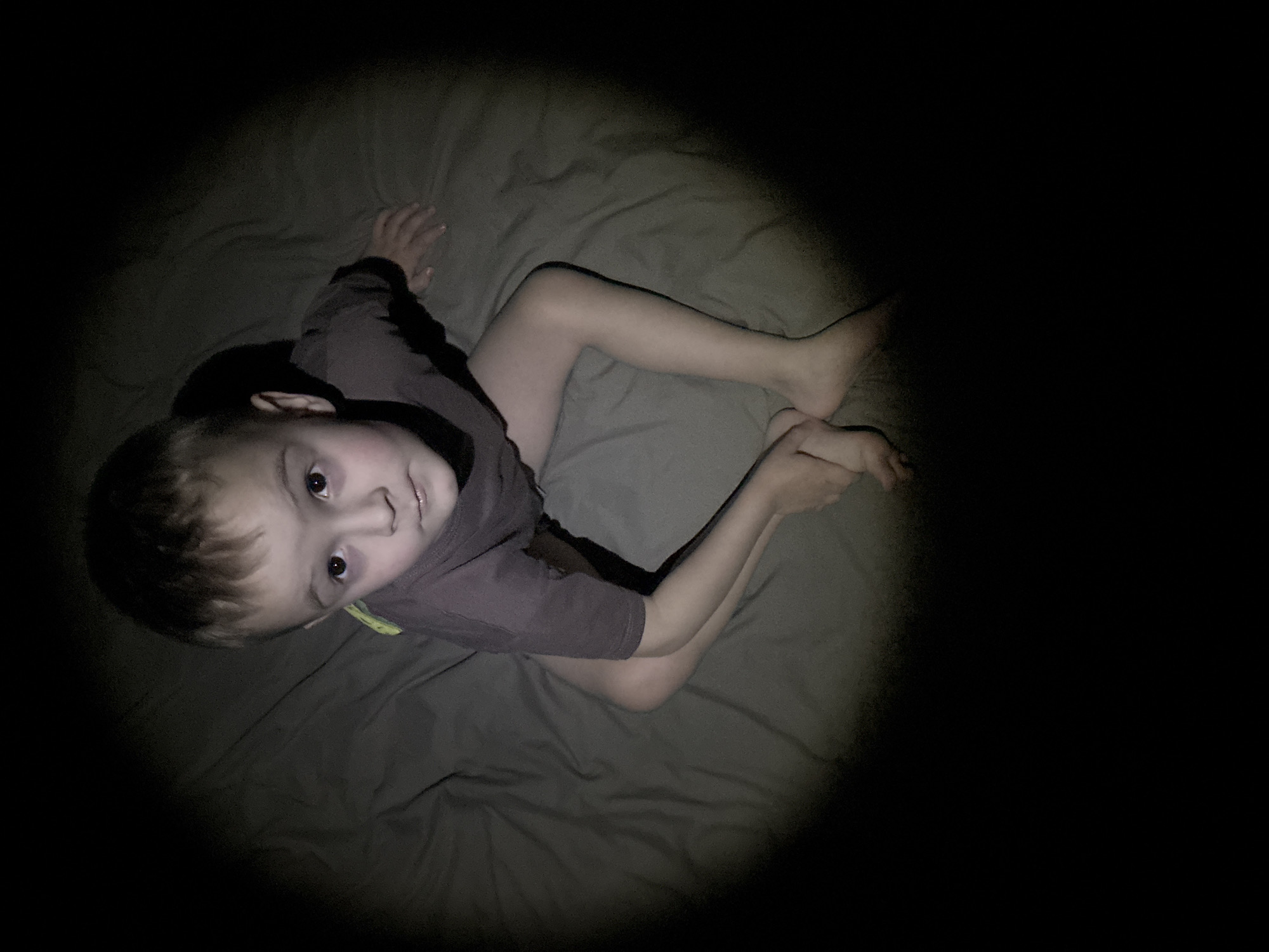A child wearing a brown, short-sleeved shirt is sitting on a grey bedsheet. The child's head is facing the camera with a slight upward gaze. The child's legs are bent at the knees and drawn up to the chest. The image is lit by a central light source, leaving the surrounding area in darkness. There is no evidence to suggest this image was taken in Chaikovsky, Russia.
