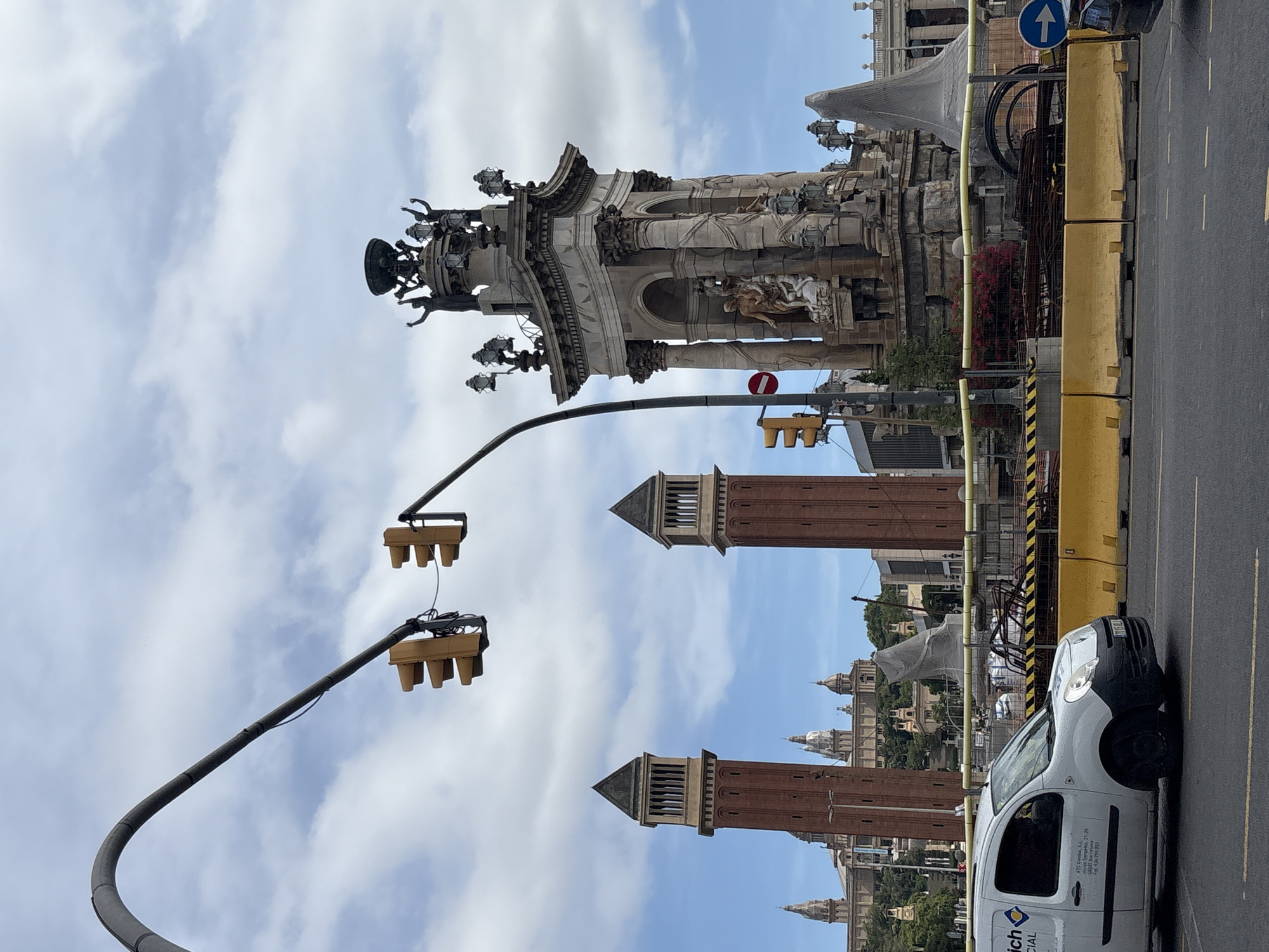 This is a street-level, eye-level, medium shot photograph taken in Barcelona, Spain, likely near the Palau Nacional on Montjuïc. The scene captures a street with traffic and a large monument.
In the foreground, a white commercial van is driving on the right side of the road. Yellow concrete barriers and construction fencing delineate a work area on the side of the road. Traffic lights are visible, with two sets hanging from a curved pole overhead. One traffic light displays red and yellow lights, while the other shows yellow lights. A circular red and white traffic sign is also present.
The middle ground features a large, ornate stone monument with sculptures and lamps. Behind the monument, two tall, red brick towers with dark, angled roofs are prominent. In the background, the grand facade of a large building with domes and arched windows, consistent with the Palau Nacional, can be seen on a hill. The sky is partly cloudy with white clouds against a blue sky. The overall impression is a busy urban environment with a historical and architectural focus.
