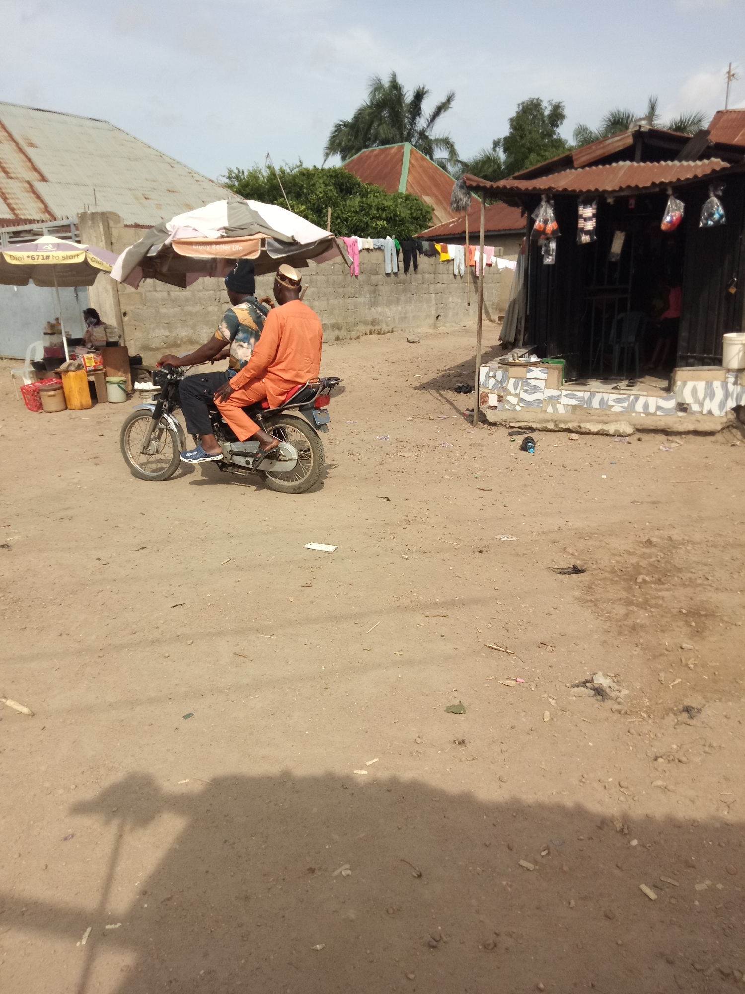 The image depicts an outdoor scene on a dirt road, possibly in a developing area. Two individuals are seated on a motorcycle. The person in front is wearing a dark and patterned shirt and black pants. The person behind is wearing an orange traditional garment and a cap. They appear to be moving. To the left of the motorcycle, there is a small roadside stall with umbrellas providing shade. Several containers and what appear to be goods are visible on the stall. Behind the stall, a corrugated metal roof of a building is visible. To the right, there is another structure with a corrugated metal roof, appearing to be a shop or dwelling. Clothes are hanging on a line in front of this structure. The ground is dusty and uneven, with scattered debris. The sky is partly cloudy.