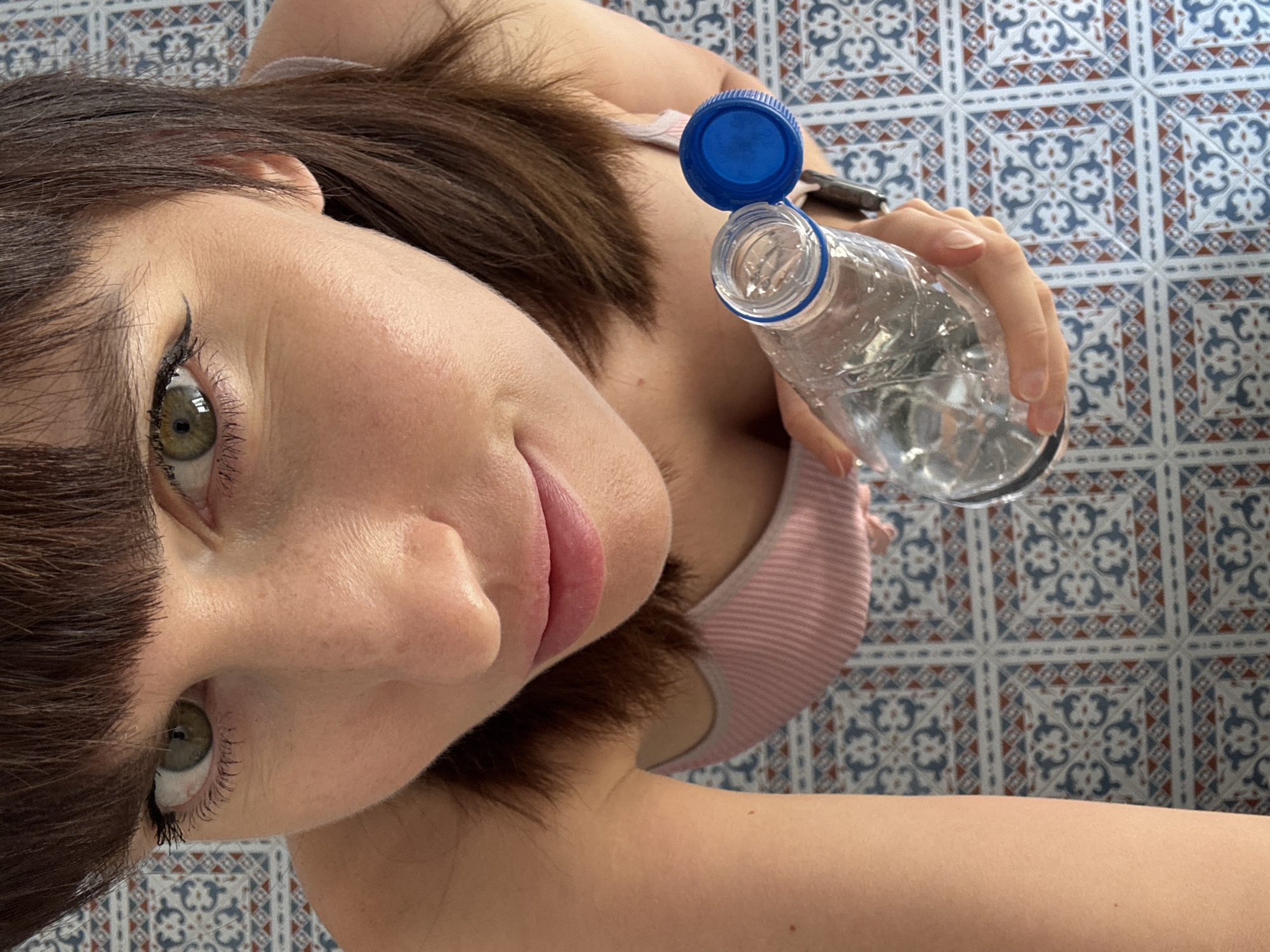 The image shows a close-up, overhead view of a woman lying down on a patterned tiled floor. She has fair skin, brown hair, and green eyes with visible eyeliner and mascara. She is wearing a pink and white striped top. Her head is tilted to the side, and her gaze is directed towards the camera. In her right hand, she holds a clear plastic water bottle with a blue cap, which is open. The water bottle appears partially full.
The background consists of a blue, white, and reddish-brown tiled surface with a repeating decorative pattern. The lighting suggests it is daytime, possibly indoors given the clear view of the tiles and the controlled lighting on the subject. The woman's expression is neutral. No text is visible in the image. The location context provided is Isola delle Femmine, Italy, which suggests the setting could be a home or a public space with this distinctive tiling.