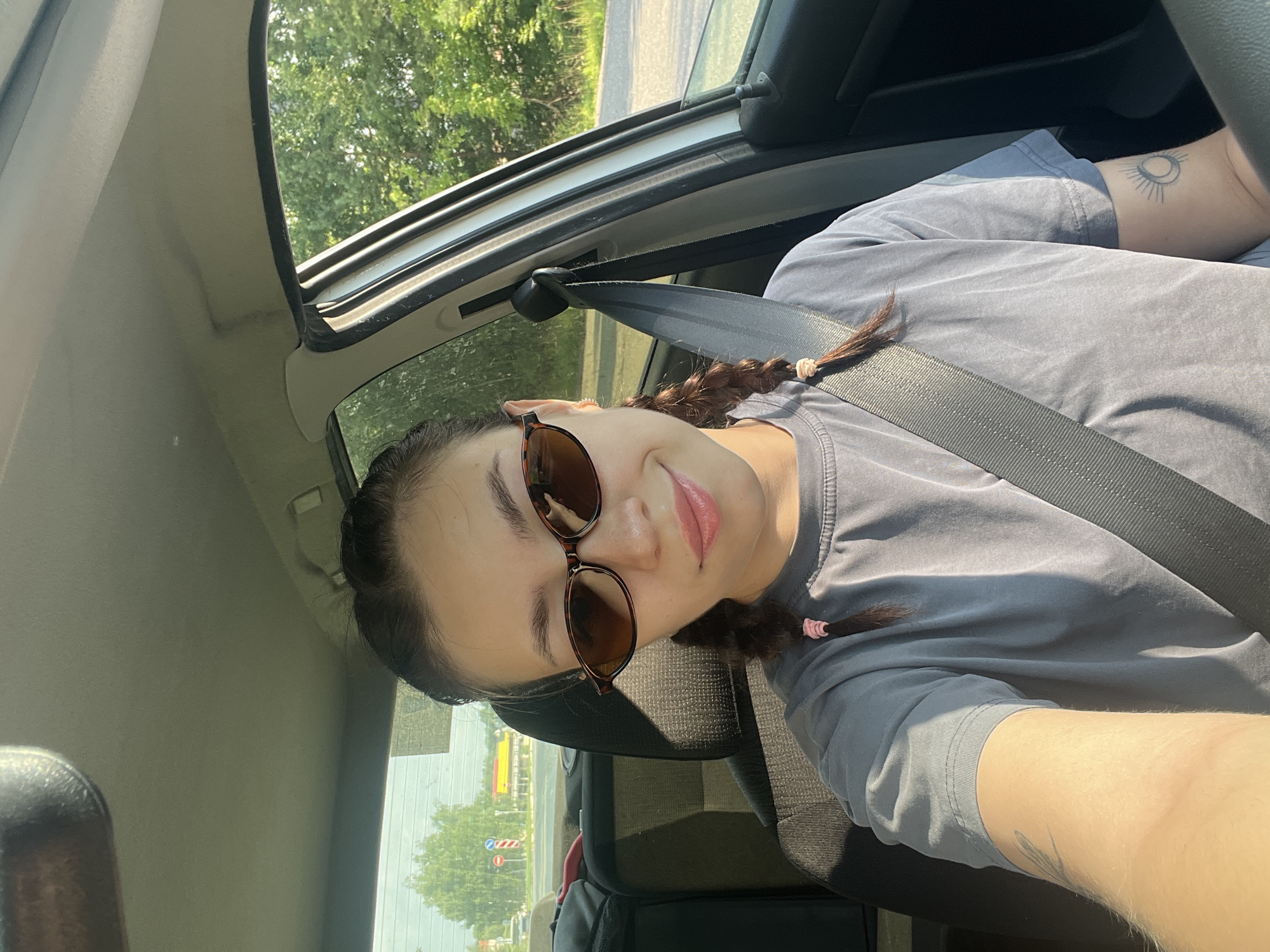 A young woman is sitting in the driver's seat of a car on a sunny day, looking at the camera. She has dark hair styled in two braids with pink ties, is wearing sunglasses, and has a seatbelt fastened across her chest. She is wearing a grey t-shirt. A tattoo of a sun and moon is visible on her left forearm. Outside the car, lush green trees and a road are visible. The scene appears to be set in or around the city of Chaikovsky, Russia, likely on a suburban or rural road given the natural scenery.
