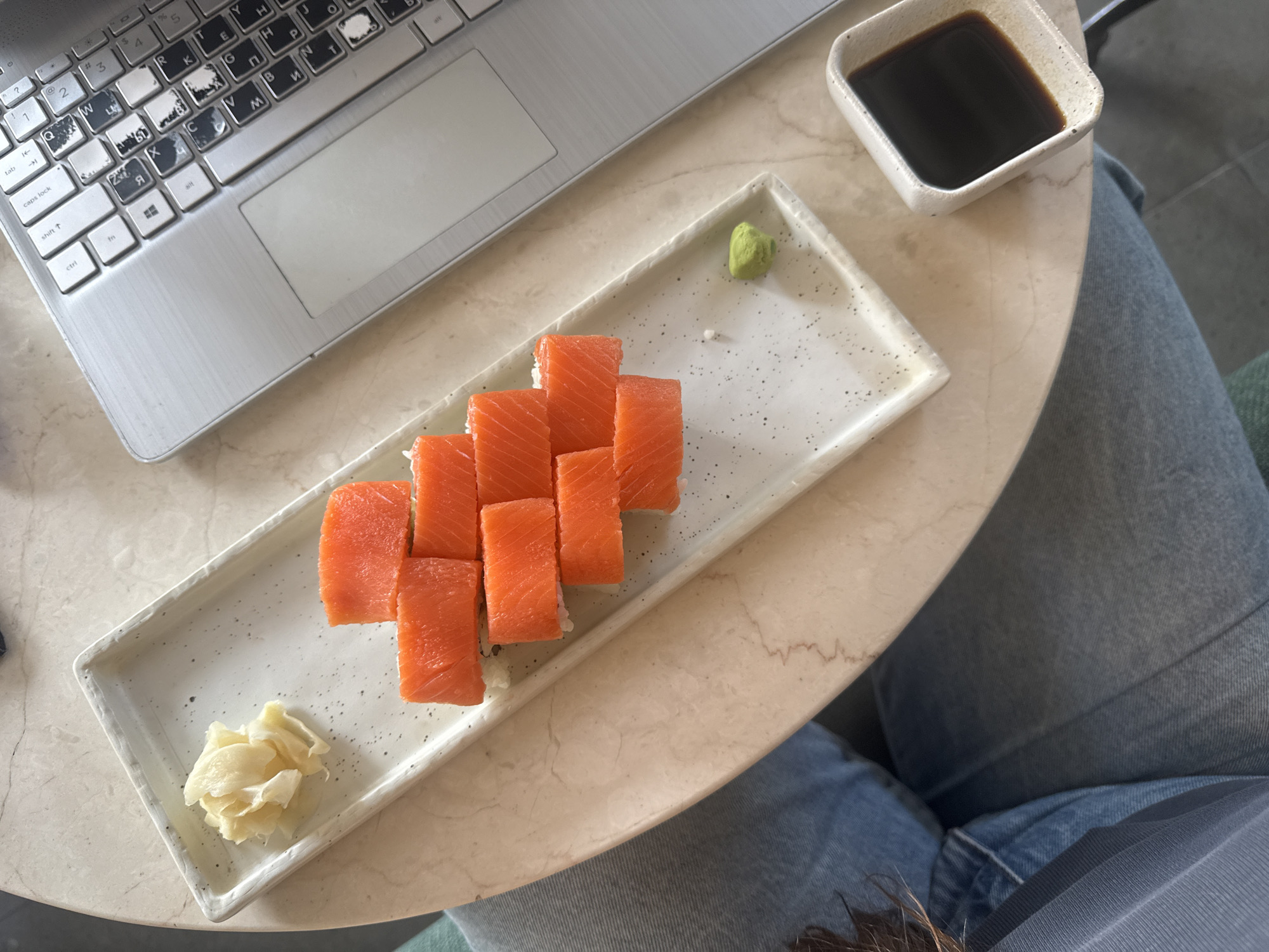 A top-down shot captures a casual dining scene in Ubud District, Indonesia, featuring a rectangular plate of sushi rolls with vibrant orange salmon, a dollop of green wasabi, and a pile of pickled ginger. A small, square white dish filled with dark soy sauce sits to the side. These food items are arranged on a light-colored, round table with a subtle marble pattern.
To the left, a silver laptop with a visible keyboard occupies a portion of the table. Many of its keys, including `Q`, `W`, `E`, `R`, `T`, `Y`, and others, show significant wear, with the black lettering worn off to reveal white underneath. The keyboard layout features both Latin and Cyrillic characters (e.g., `Я`, `И`, `Т`, `П`). Visible key labels include `tab`, `caps lock`, `shift`, `ctrl`, `fn`, and `alt`, along with numbers `1` through `5` and symbols like `?`, `@`, `#`, `$`, `%`.
Part of a person's lap, clad in blue denim jeans, is visible on the right side and bottom of the frame, suggesting they are seated at the table. The lighting indicates it is daytime. The scene portrays someone enjoying sushi, possibly while working or relaxing with their laptop nearby.