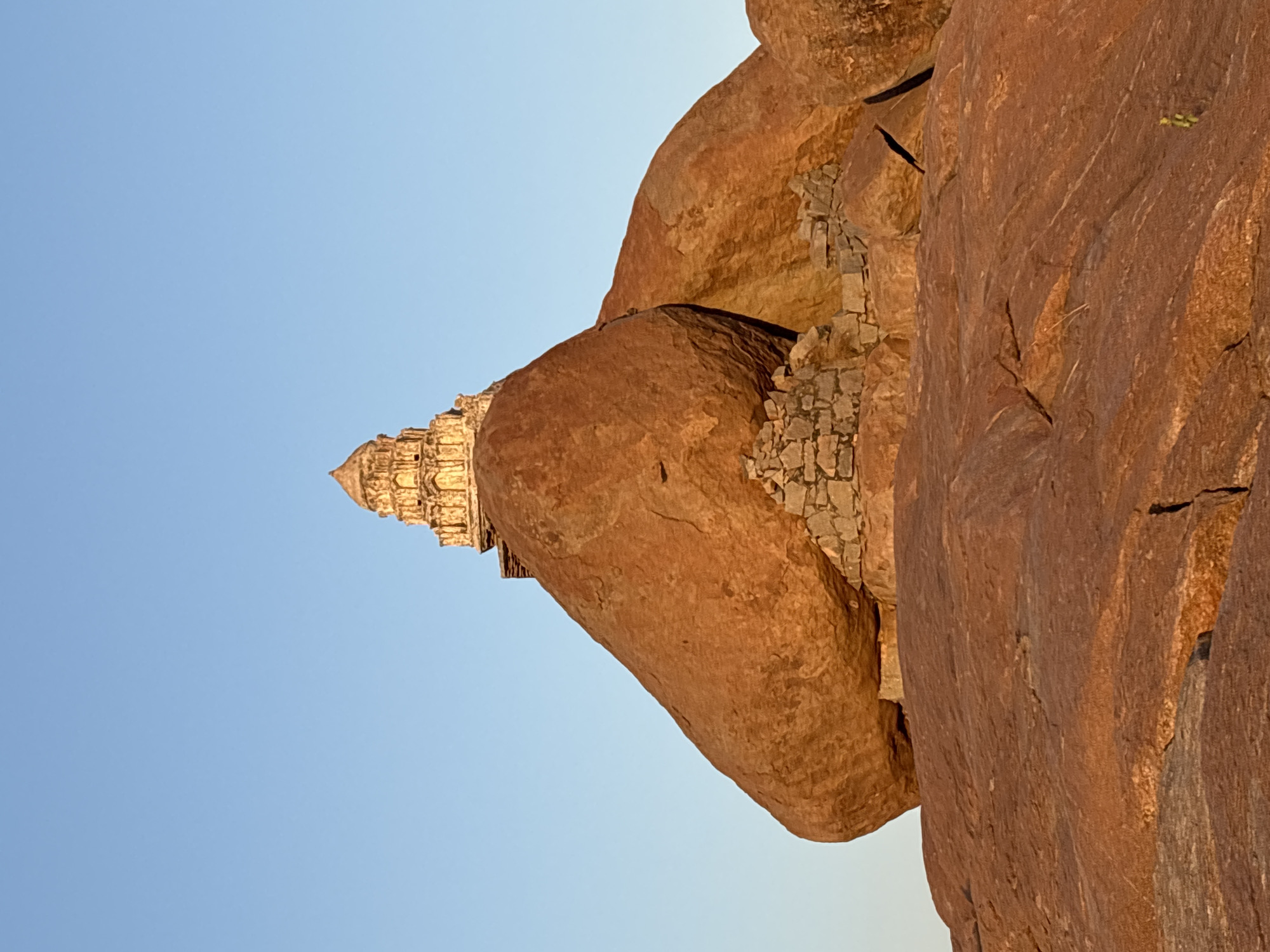 The image shows the Achyutaraya Temple, a ruined temple complex in Hampi, near Kamalapura, India. The temple is partially obscured by large, reddish-brown boulders typical of the Hampi landscape. A small portion of the blue sky is visible in the upper left corner of the image. The temple itself is a light color and intricately carved. Smaller stones appear to be wedged between the boulders.