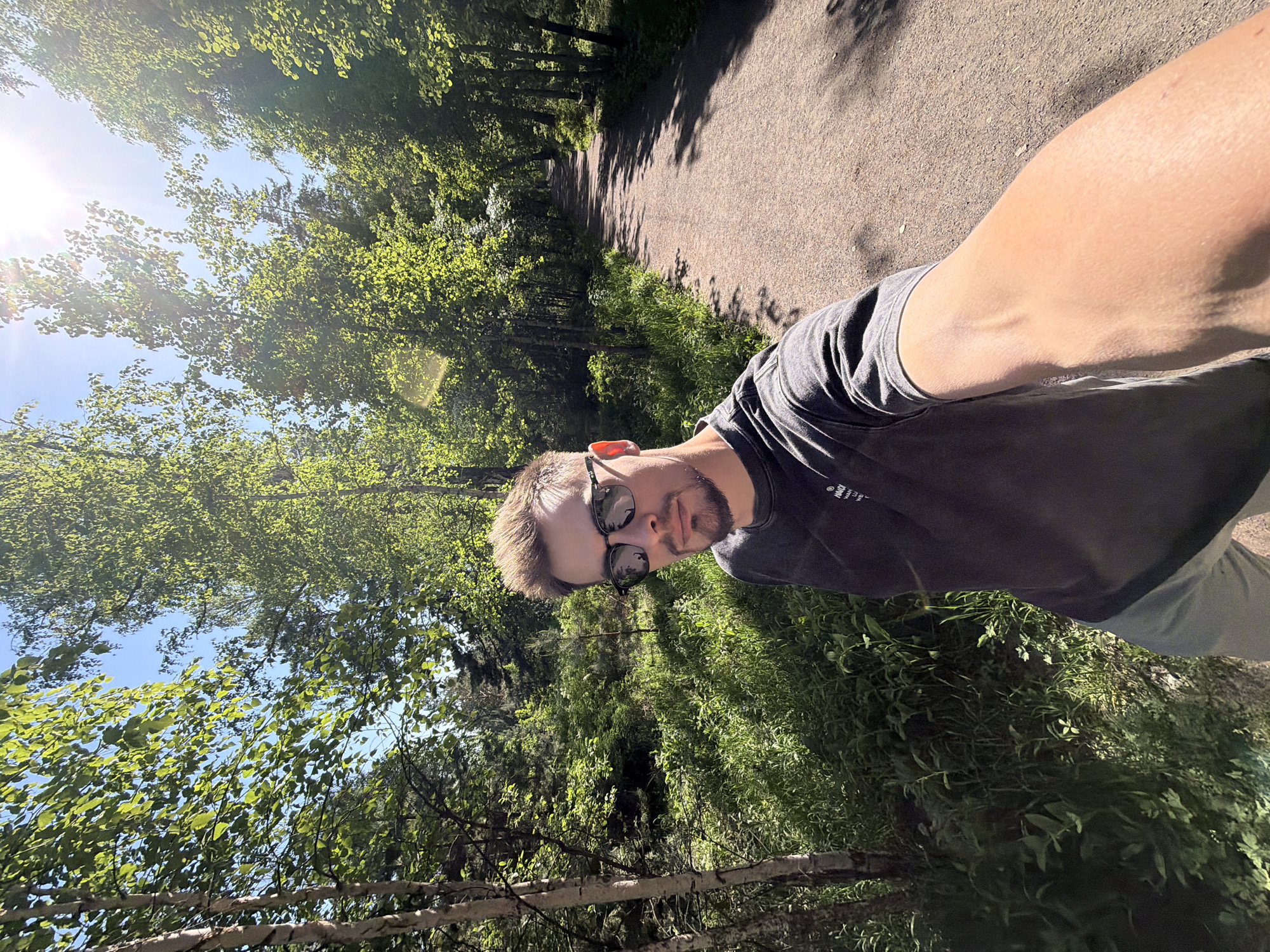 This media file is a selfie taken by a man on a sunny day in what appears to be a park or forest path in Krasnoyarsk, Russia. The man is positioned in the lower right foreground, looking directly at the camera. He has short, light brown hair, a neatly trimmed beard, and is wearing dark sunglasses and a dark gray t-shirt. On the left sleeve of his shirt, partially visible white text appears to begin with "HMOI" or "HMOT".
The background features a tree-lined path that recedes into the distance. Lush green foliage and trees dominate the scene, suggesting late spring or summer. Bright sunlight streams through the tree canopy from the left, creating visible lens flare and sunbeams in the upper left portion of the image. Long shadows from the trees are cast across the dirt or gravel path, indicating either morning or late afternoon. The sky is bright blue and clear. The overall impression is one of a pleasant, vibrant outdoor environment.
