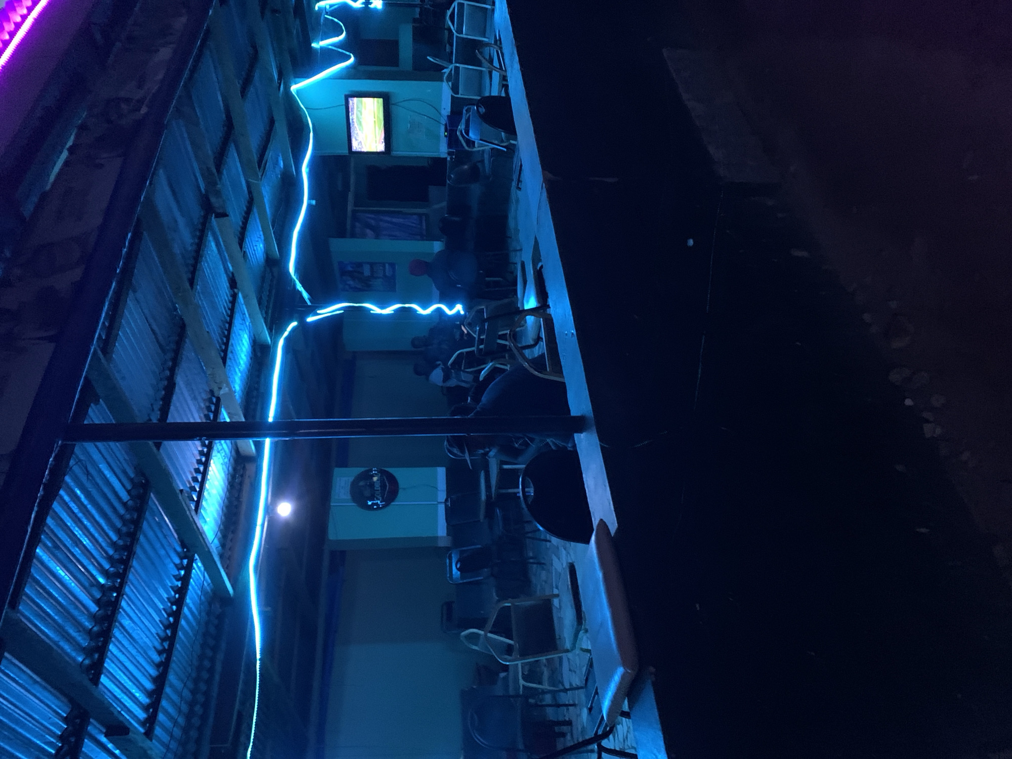 The scene depicts the interior of an internet cafe located in New Stadium, Nigeria, at night. The cafe is dimly lit with blue neon lights running along the corrugated metal roof and the wall. Several individuals, possibly customers, are seated at computers. Some of these individuals have their backs turned to the camera. One individual is wearing a red hat. Several computer stations are visible. A small television is mounted on the wall, displaying a green image. A circular sign with the word "GOLDEN" on it is also visible. The floor appears to be tiled or concrete. The right side of the image is black.