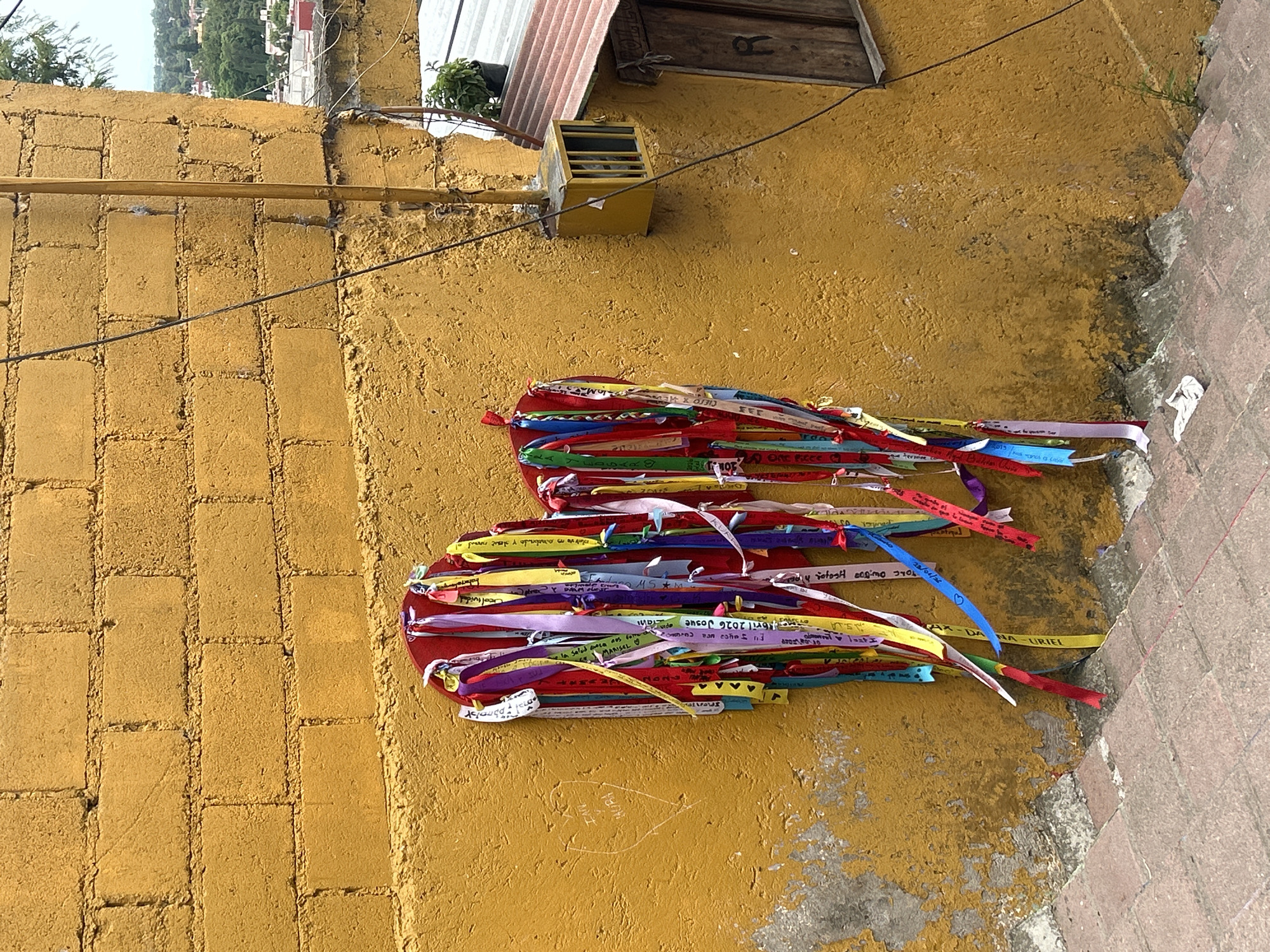 The image captures a vibrantly yellow, rough-textured brick wall in Atlixco, Mexico. A heart-shaped structure, made of red material and adorned with numerous colorful ribbons, hangs from the wall. These ribbons, inscribed with various messages in Spanish, are meant to represent wishes or prayers. To the left of the heart, faint markings in the yellow paint suggest a carved heart with text inside. A stone staircase, made of reddish-brown bricks, leads downwards in the foreground, with some debris scattered on the steps. To the right of the heart, a small, yellow utility box with a caged opening is attached to the wall, and further to the right, a wooden window with the letter 'R' carved into it is visible. The background shows a glimpse of a green landscape and buildings, indicating an urban environment. The overall scene depicts a place of cultural and personal expression within the city.