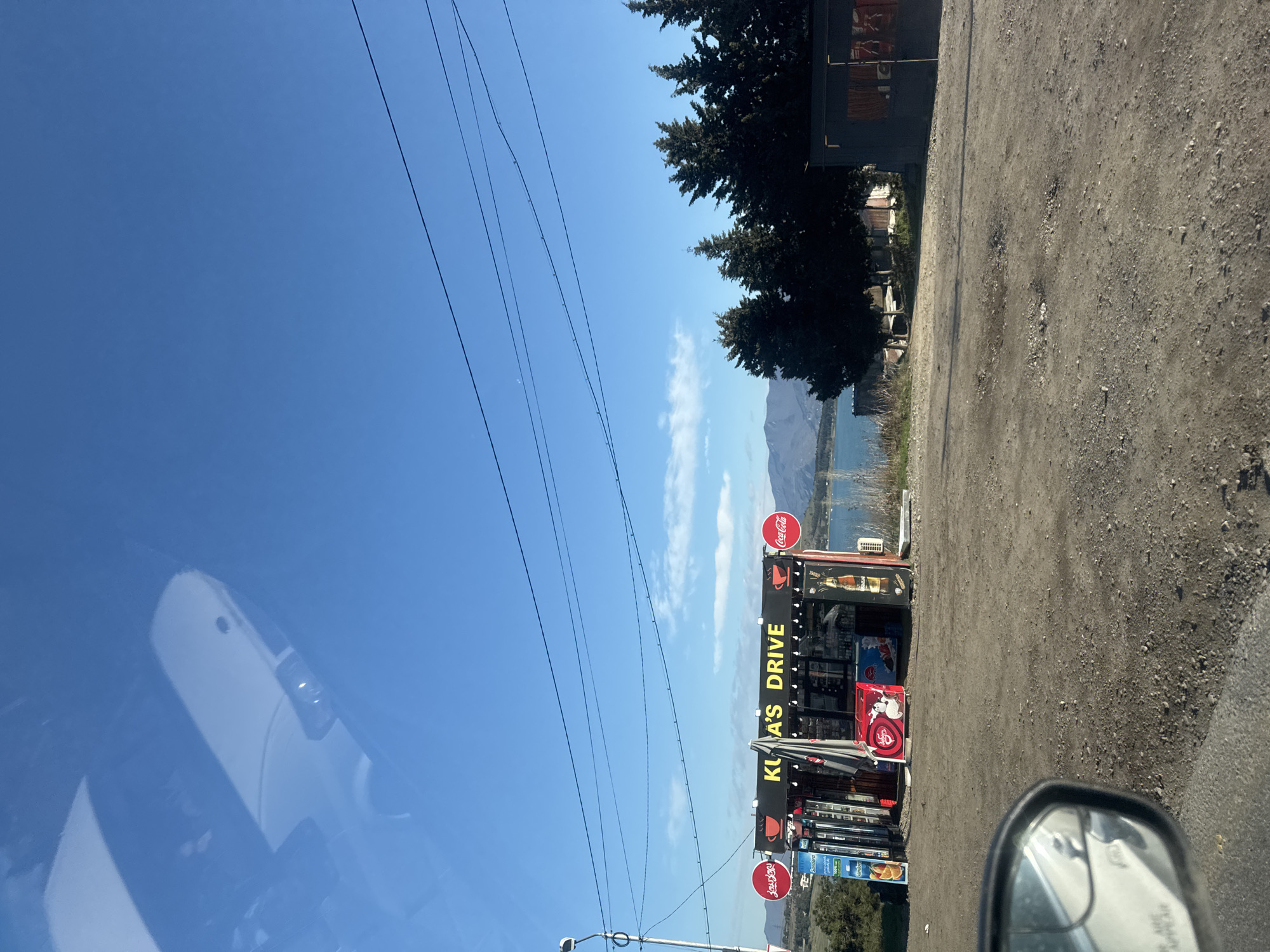 The image shows a roadside view from a car, likely in the vicinity of Tbilisi, Georgia, based on the presence of "Kura's Drive," a known establishment in the region. The sky is clear and blue, with power lines running across the frame. To the right is a gravel road, leading to a drive-in style shop identified as "Kura's Drive." Coca-Cola signs are visible. Further back, there are trees, water (possibly a lake or river), and a mountain in the distance. A portion of the car's side mirror and windshield are visible in the foreground.