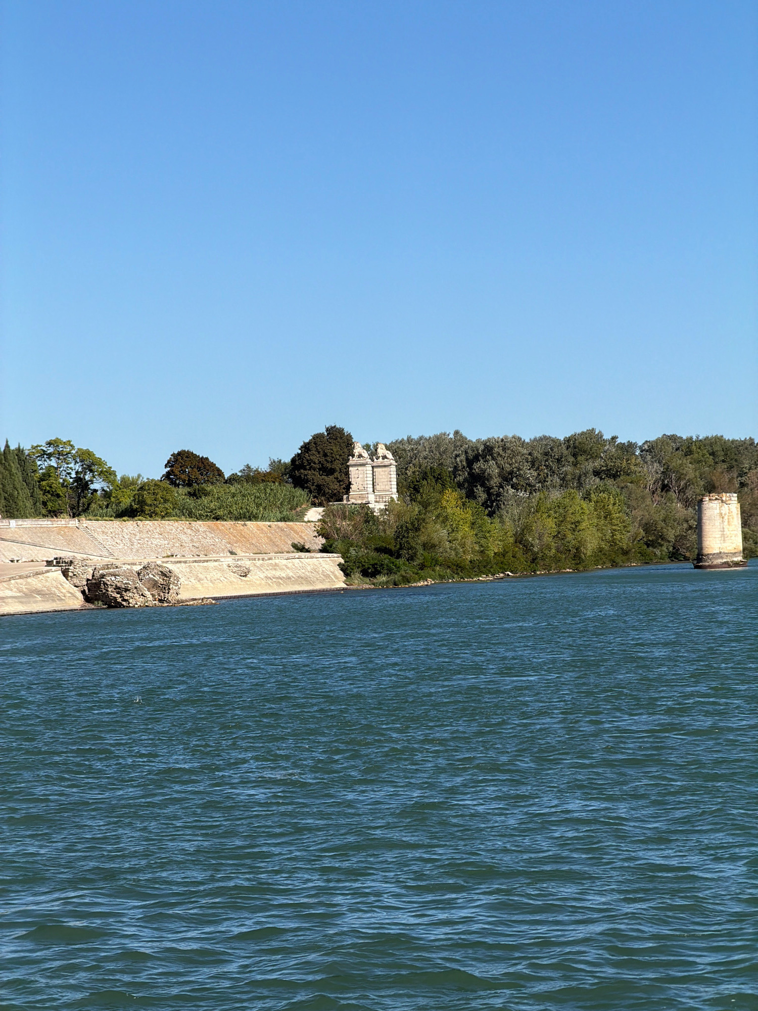 The image shows the Rhône River in Arles, France. On the far bank, there is a stone embankment with some ruins. Above the embankment, there are trees and a building with classical architecture, possibly part of the Roman theatre or amphitheater. In the river, there is a lone stone pillar, likely a remnant of an ancient bridge. The water is blue and rippling, reflecting the clear blue sky. The scene is tranquil and historic, evoking the ancient past of Arles.