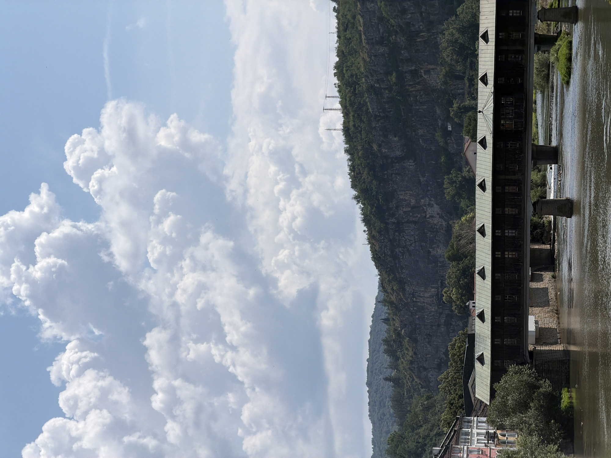 The image shows the covered bridge of Lovech, Bulgaria, spanning the Osam River. The bridge is a wooden structure with a green roof and triangular dormer windows. On the left side of the bridge, there are buildings with pink and white facades. The river is calm and reflects the sky, which is filled with large, fluffy white clouds. Behind the bridge and river, there is a rocky cliff face covered with lush green vegetation and trees. Power lines and transmission towers are visible on the ridge of the cliff. The overall scene depicts a picturesque and historic urban landscape with natural elements.