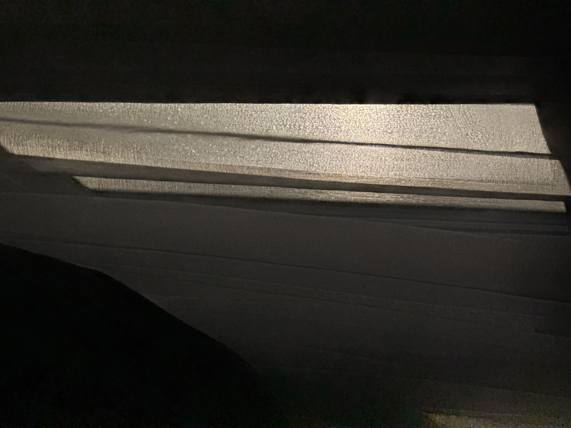 This is a photograph of a dark room with a lit curtain. The curtain is a sheer, white fabric that is illuminated from behind, casting shadows and creating vertical lines of light and dark. The lighting suggests that there may be a window or light source behind the curtain. The room itself appears to be largely in shadow, with a dark, indistinct mass on the left side of the image. The overall impression is one of mystery and perhaps confinement. The location of Doma 1096 Km, Russia, does not provide any additional context to the image.