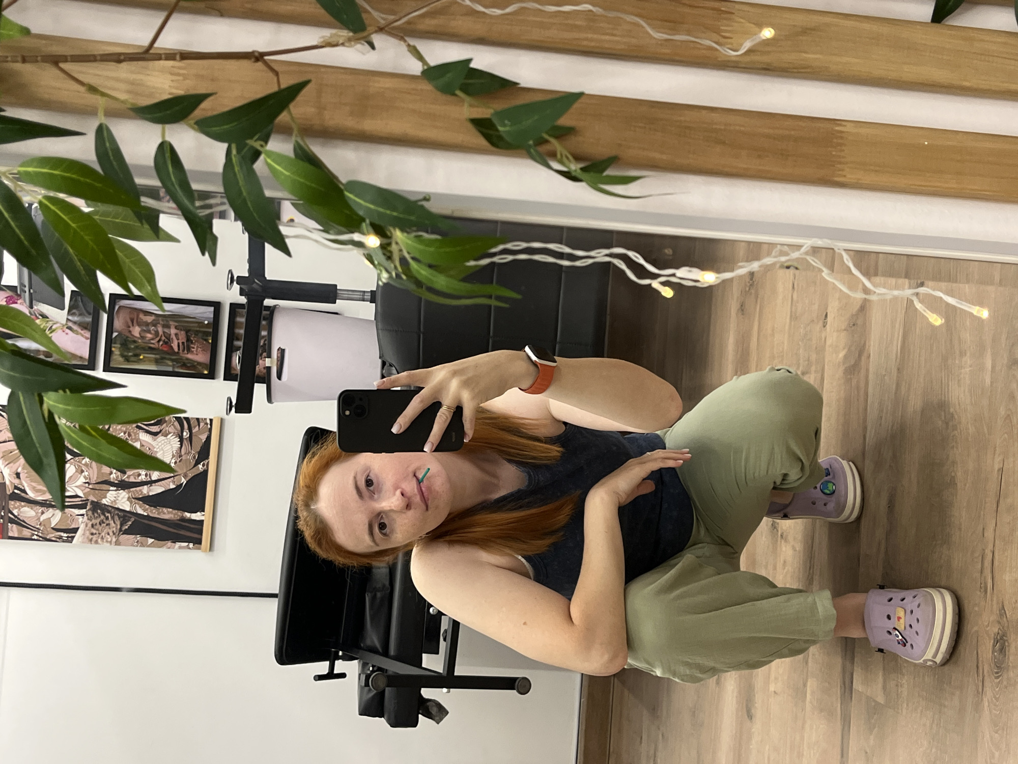 A woman with red hair, wearing a dark blue tank top, light green pants, and purple Crocs, is captured in a mirror selfie. She is holding a smartphone in her right hand and has a green object, possibly a toothpick or a small decorative item, in her mouth. She is crouching on a wooden floor in what appears to be an indoor space. In the background, a black tattoo chair and framed artwork are visible. To the right of the frame, a plant with green leaves and fairy lights are present, along with wooden vertical panels. The setting is likely a tattoo studio or a similar establishment. There is no information in the image that can place the location in Chaikovsky, Russia.