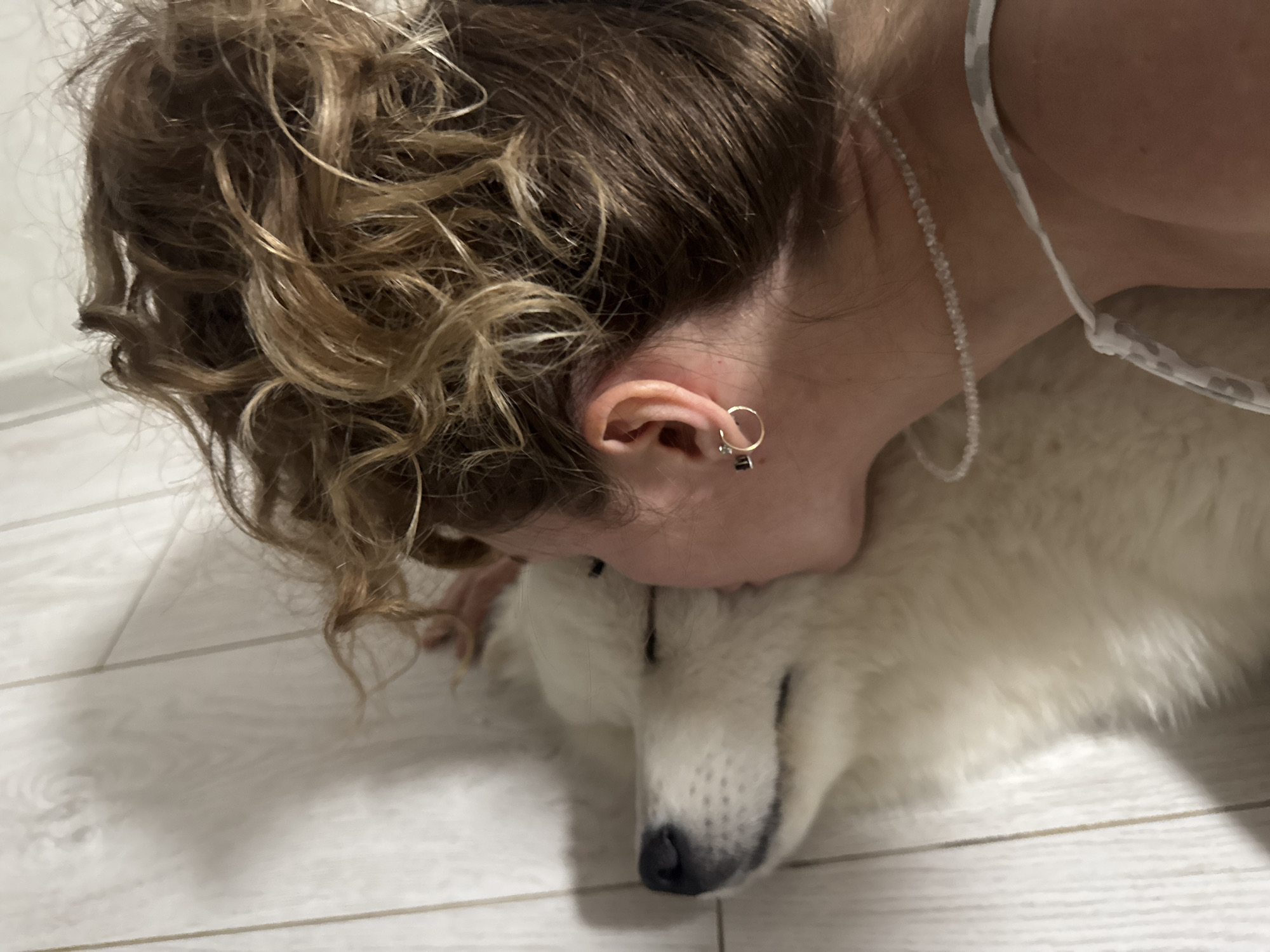 A close-up, low-angle shot shows a person with curly brown hair lying down, their face nuzzled against the head of a white dog. The person is wearing a necklace and an earring. The dog's eyes are closed, and its head is resting on a light-colored wooden floor. The background is out of focus, showing a hint of white wall. The scene appears to be indoors, potentially in a home.