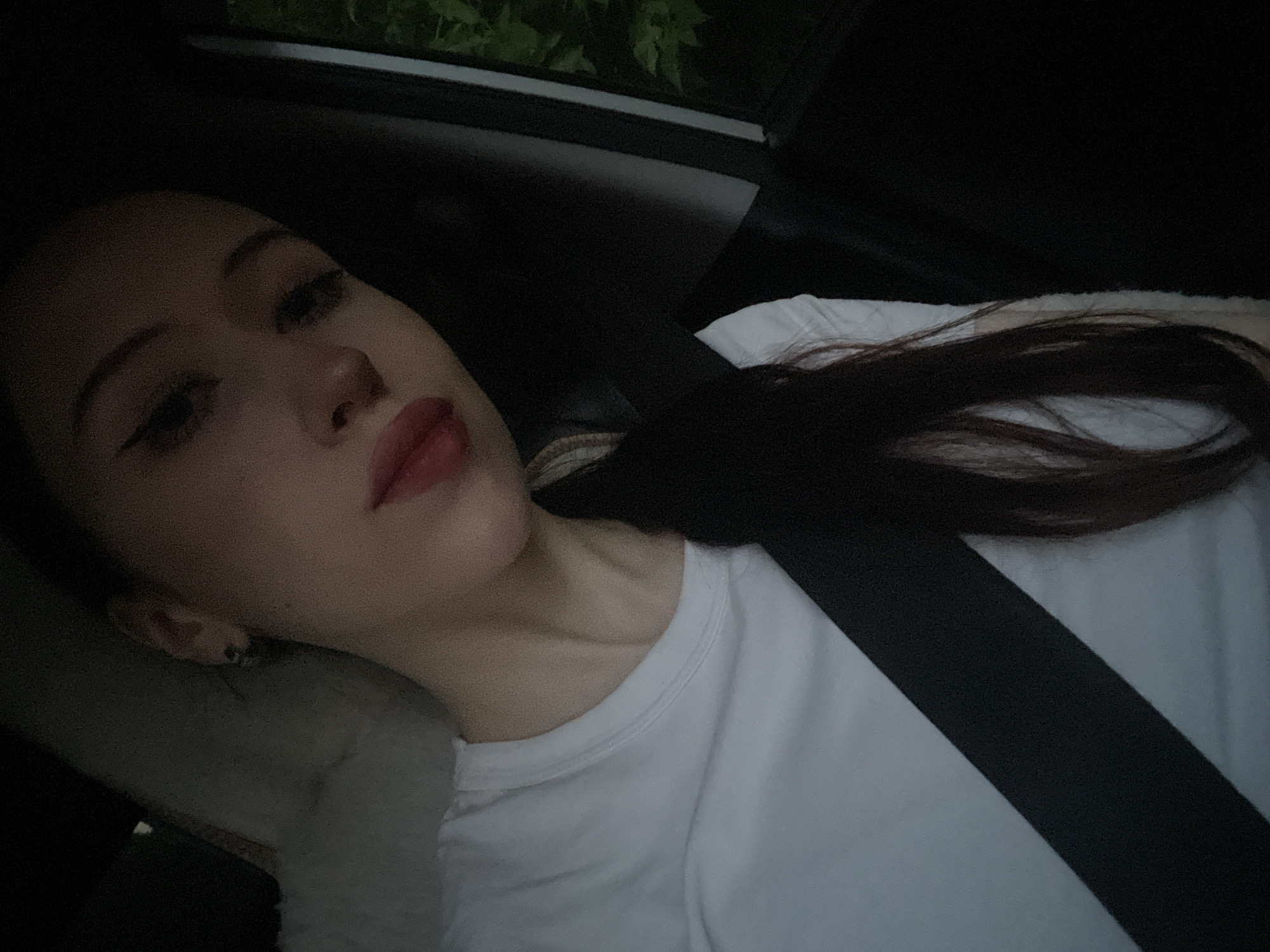 The image shows a young person wearing a white t-shirt and a black seatbelt, sitting in what appears to be a car. The person has dark hair styled in a braid, and is wearing makeup, including winged eyeliner and red lipstick. The background is dark, with some greenery visible outside a window. The overall lighting suggests it might be dusk or evening. There are no specific landmarks or details in the image that would allow for identification of the location as Votkinsk, Russia.