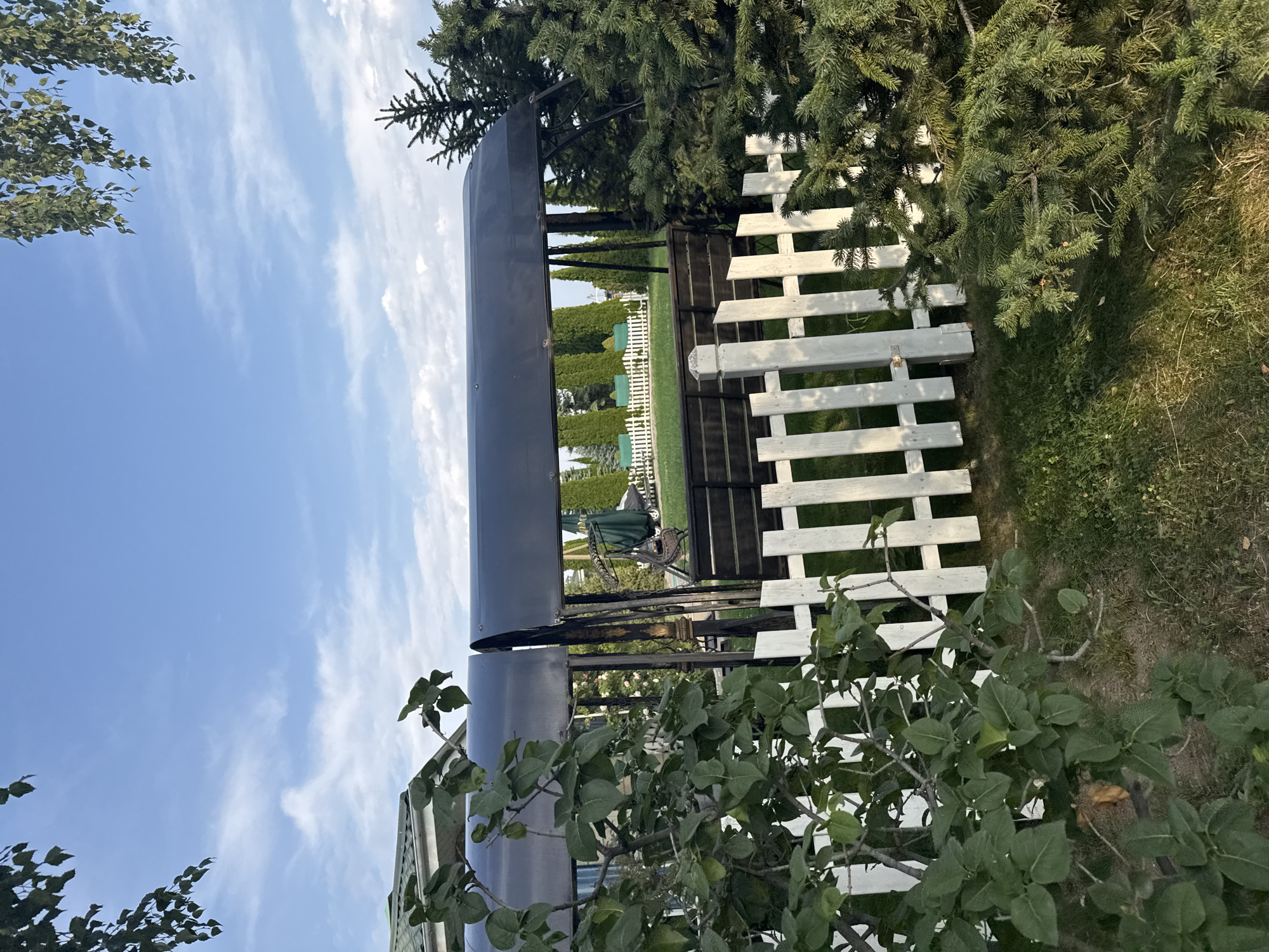 The image depicts a backyard scene in Sary-Oy, Kyrgyzstan, under a partly cloudy blue sky. In the foreground, a white picket fence runs across the lower portion of the frame, partially obscured by lush green foliage. Behind the fence, a large, dark-colored swing set with a canopy is visible. The swing set has a dark metal frame and a dark fabric canopy. Further in the background, manicured green grass is visible, with several rows of tall, slender green trees lining a white fence in the distance. Some evergreen trees are also present on the right side of the image. The overall impression is that of a well-maintained outdoor recreational area.