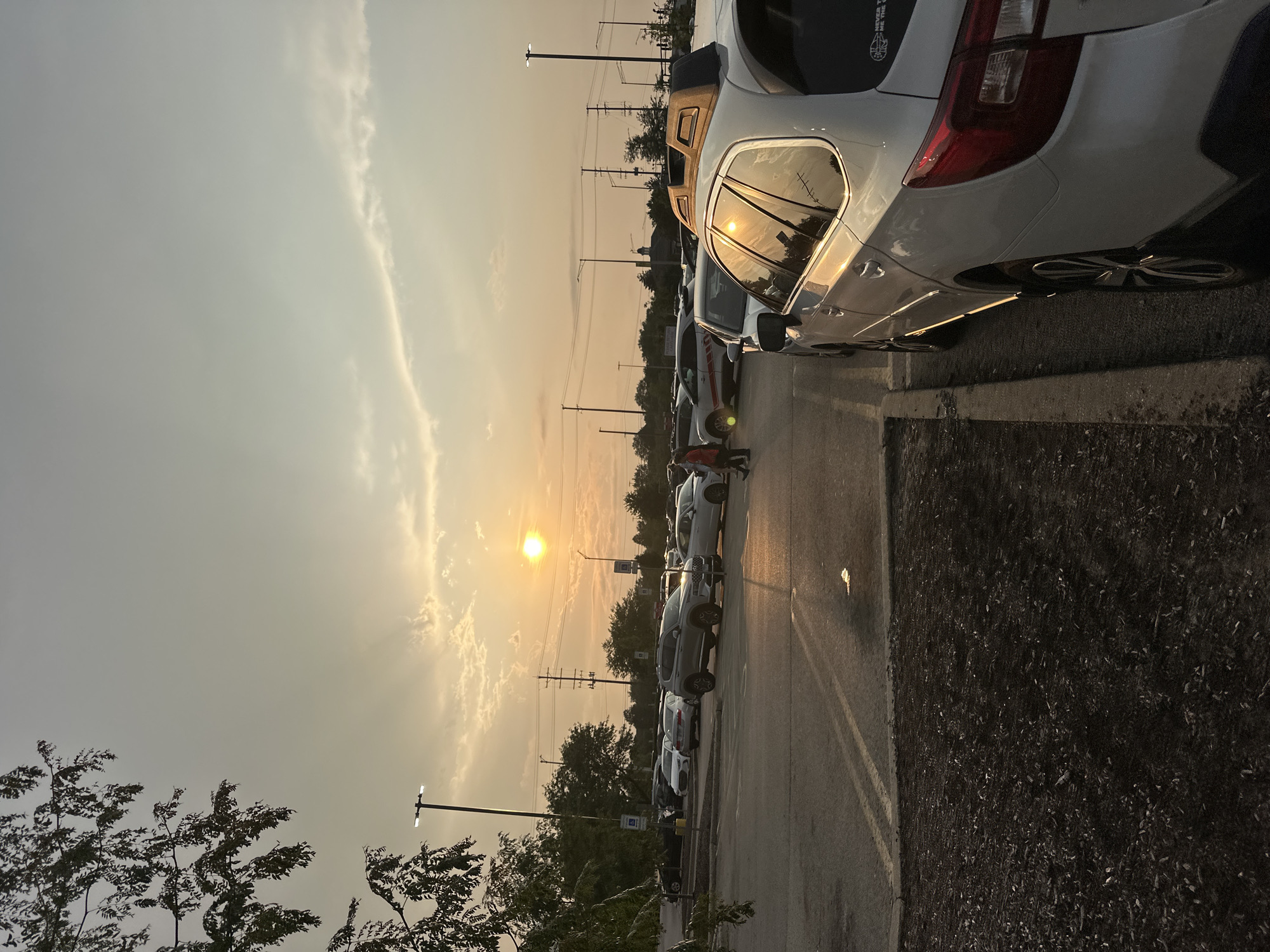 A sun-drenched outdoor parking lot is shown at sunset. The sun is visible as a bright, hazy circle in the sky, casting a warm glow on the scene. Several cars are parked in rows, with a white Subaru Outback in the foreground to the right. A person in a red shirt and dark pants is walking in the middle distance. The parking lot is paved, with a dirt and mulch area to the left. Trees and utility poles are visible in the background, and power lines stretch across the sky. The overall impression is one of a typical suburban commercial area at the end of the day.