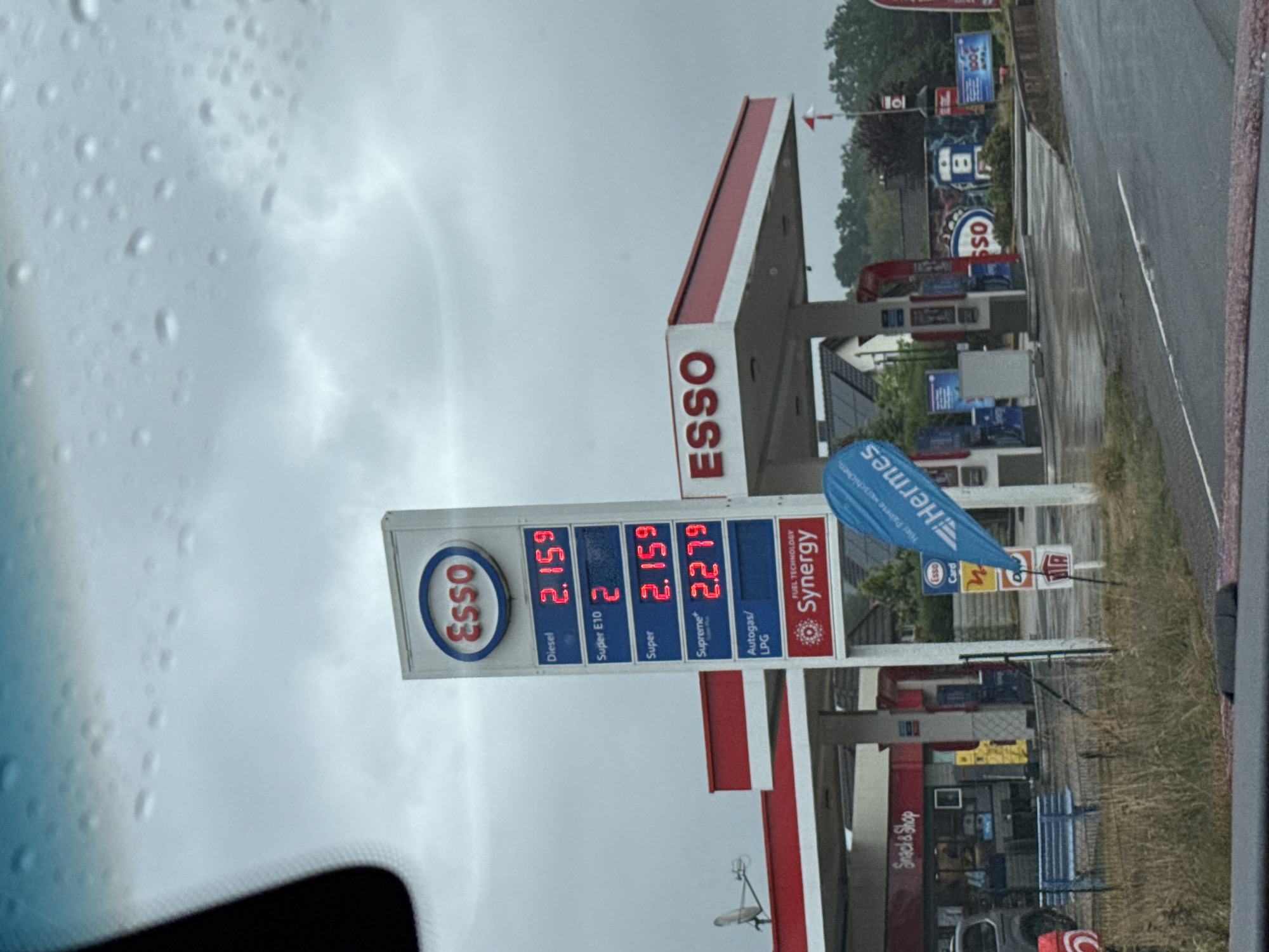 The image shows a gas station on a cloudy day. The gas station has a large red and white canopy, with an ESSO sign at the top. A price board displays the cost of different types of fuel: Diesel at 2.159, Super E10 at 2, Super at 2.159, and Supreme at 2.279. Below the fuel prices, there is a FUEL TECHNOLOGY Synergy logo. There are also various signs visible, including one for "Snack & Shop" and a blue flag with the ESSO logo and text that partially reads "Hier tanken ...". In the background, there are houses, trees, and other gas pumps. The ground is wet, indicating recent rain. A car is parked at the gas station. The scene appears to be located at an ESSO gas station in the city of Bad Sachsa, Germany.