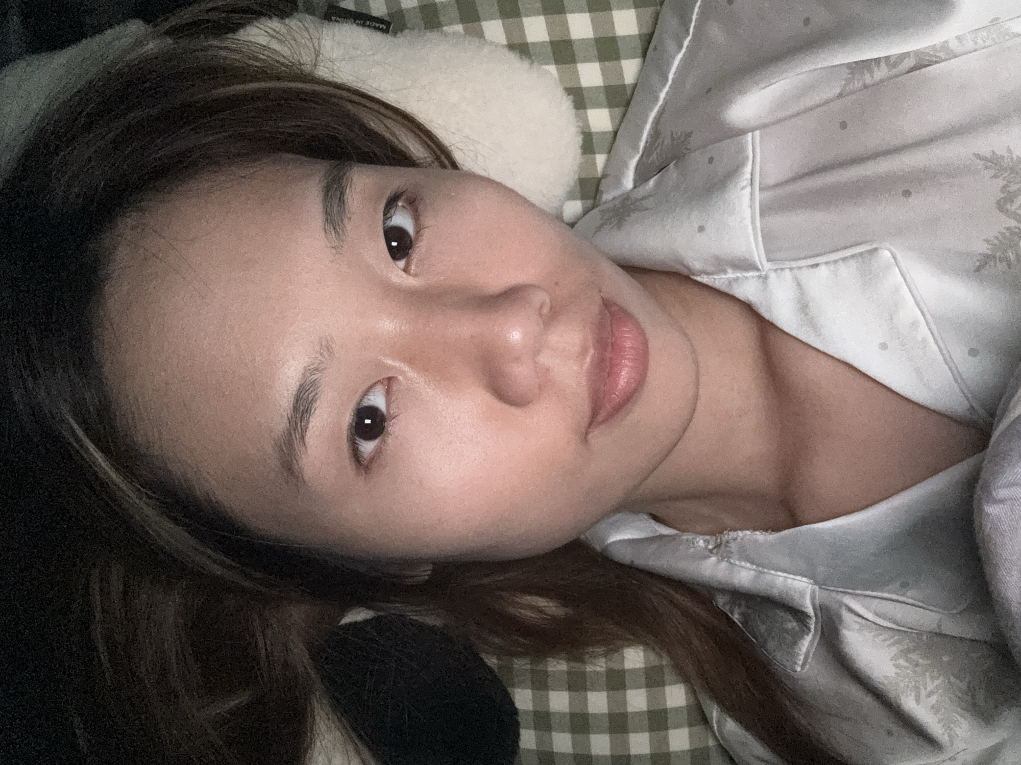 A woman lies on a bed. She is on a pillow with a white, fluffy cover. She is wearing light-colored patterned pajamas with a collar. The bed has a green and white checkered pattern. The location of the photograph is unknown.