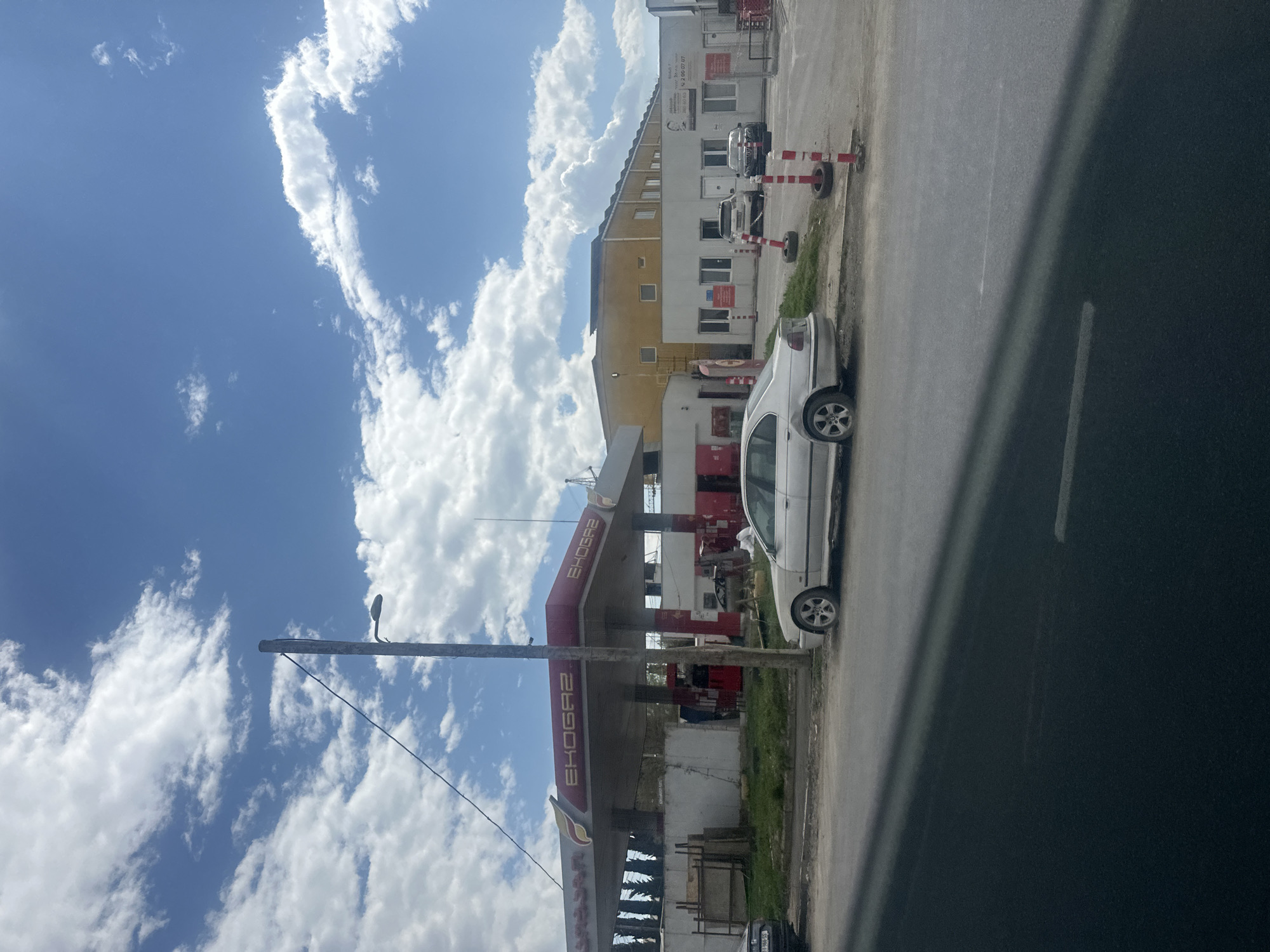 The image shows a daytime scene in Tbilisi, Georgia. An EkoGaz gas station is visible. A light-colored car is parked on the side of the road next to the station. In the background, a white and yellow building is partially obscured by the clouds. A utility pole is visible in the foreground. The sky is blue with scattered white clouds. Other vehicles and traffic cones are visible near the building in the background. The image is taken from within a vehicle.