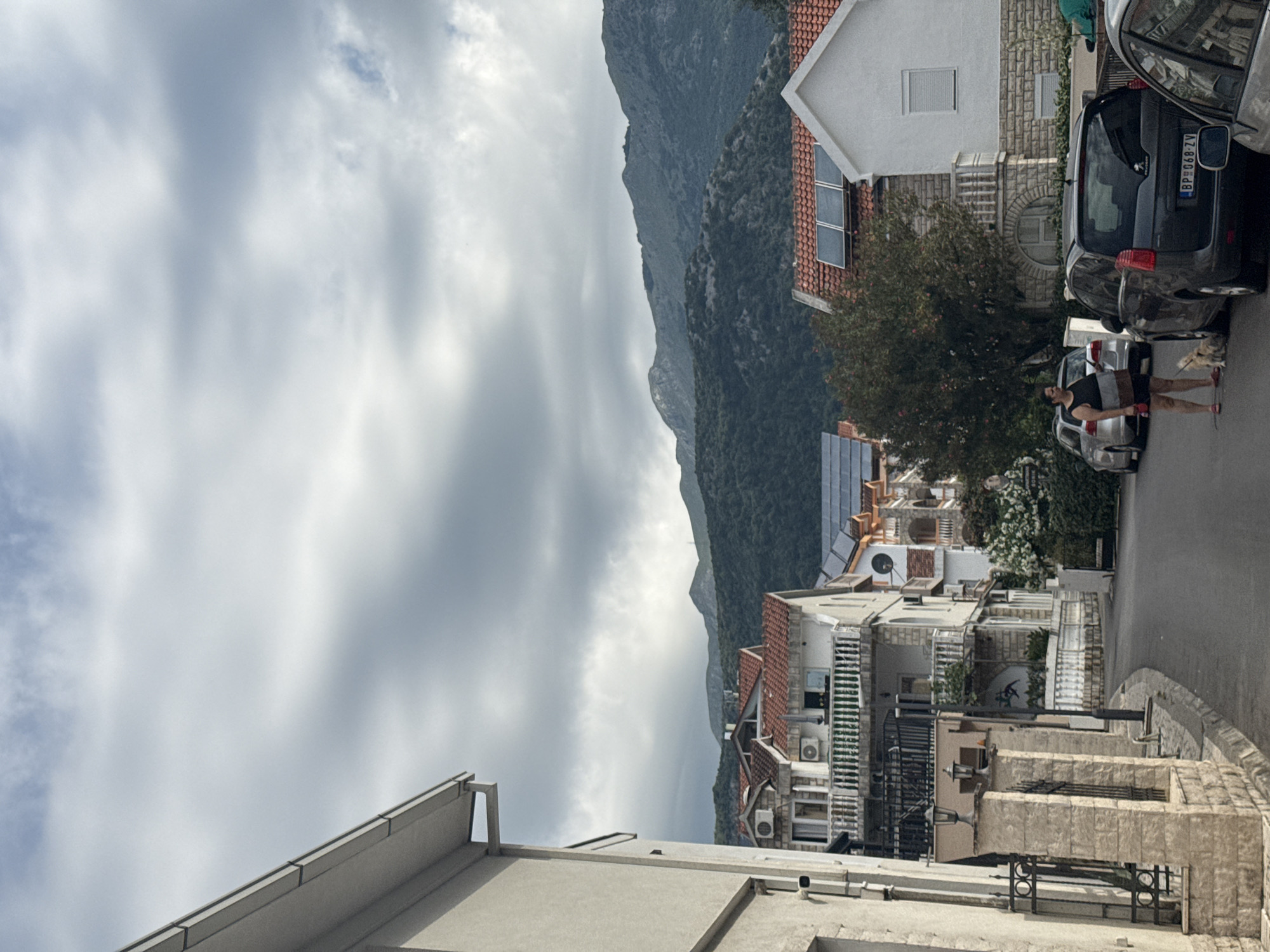 The image displays a vibrant residential street scene in Budva, Montenegro, set against a backdrop of imposing mountains. The perspective looks up a gently sloping street.
In the mid-ground, a paved road stretches, lined with several parked cars. A person, appearing to be a woman, is visible standing next to a silver car, accompanied by a small, light-colored dog. She wears a dark top, light shorts, and striking red shoes, seemingly pausing or waiting.
To the left of the street, a collection of multi-story houses and apartment buildings rise. Many structures feature traditional red tile roofs, while others have modern flat roofs. Several roofs are equipped with solar panels, and numerous air conditioning units are affixed to building exteriors. Stone facades and fences are prominent, contributing to the local architectural style. A lush green tree stands between the street and the buildings.
The background is dominated by towering, steep mountains covered in dense, dark green vegetation, characteristic of Montenegro's rugged coastal landscape. The sky above is largely overcast with soft, diffuse light, suggesting a daytime scene under cloudy conditions. The atmosphere appears calm. A visible Montenegrin license plate on one of the parked cars reads "BP 068-ZV".