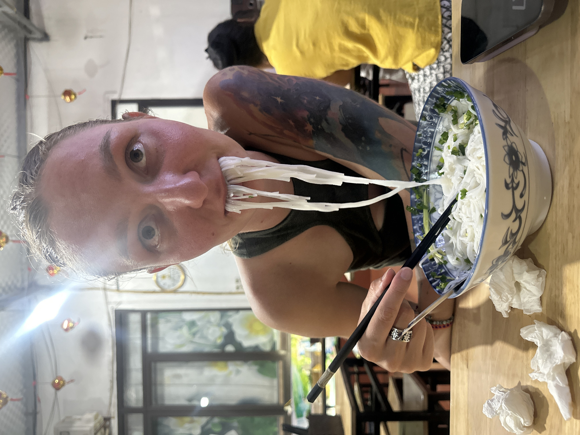 The image depicts an indoor restaurant scene in Trung Tam Ward, Vietnam. The central subject is a fair-skinned individual with light hair, seen from the chest up, positioned directly in front of the camera, looking straight ahead with wide eyes. This individual is actively consuming white rice noodles from a blue and white patterned ceramic bowl, with a significant portion of noodles hanging from their open mouth. They hold black chopsticks in their right hand, with a silver-colored ring on the ring finger. A large, multi-colored tattoo, depicting a celestial or cosmic theme with blues, purples, and oranges, covers their left arm. They wear a dark tank top.
The bowl of noodles contains chopped green onions, and a silver spoon or utensil rests inside it. On the light-colored wooden table in front of the bowl are three crumpled white paper napkins or tissues. To the right of the bowl, a dark-colored smartphone lies on the table.
In the background, another individual wearing a bright yellow t-shirt is partially visible on the right, seated at a table. The restaurant environment features white walls, with several red and gold decorative lanterns hanging from the ceiling. An air conditioning unit is mounted on the upper left wall. A window or framed artwork with a floral design is visible through a doorway in the middle-left background. Another circular decorative object is present on the right wall. The scene is brightly lit, with a strong light source visible at the top, creating glare and highlights.