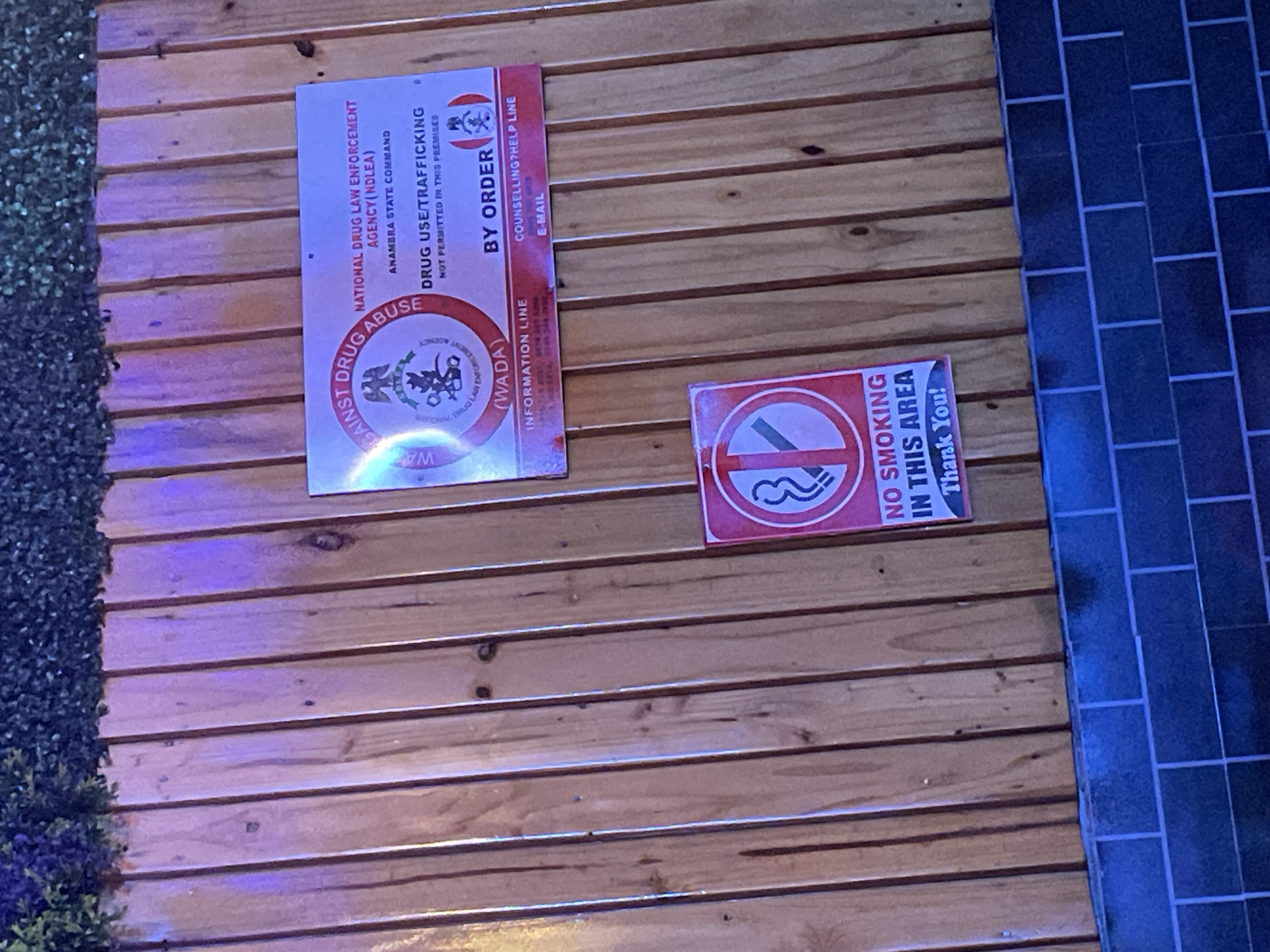 The image displays a section of a wall within an establishment in Obosi, Nigeria, featuring two regulatory signs. The wall is predominantly covered with horizontal wooden paneling, flanked by artificial greenery on the far left and dark blue/black square tiles on the far right. The lighting suggests an indoor setting, possibly with artificial illumination creating a slight purple hue.
Two signs are mounted on the wooden wall. The larger sign on the left is a public awareness notice from a drug enforcement agency. It clearly states "NATIONAL DRUG LAW ENFORCEMENT AGENCY (NDLEA)" and "ANAMBRA STATE COMMAND." A prominent warning reads, "DRUG USE/TRAFFICKING NOT PERMITTED IN THIS PREMISES," followed by "BY ORDER." Below, there are sections for an "INFORMATION LINE" (associated with "(WADA)") and a "COUNSELLING?HELP LINE" with an "E-MAIL" field, though specific contact details are not fully legible. The top banner of this sign declares "AGAINST DRUG ABUSE" and includes a logo of the enforcement agency.
The smaller sign on the right is a standard "NO SMOKING" notice. It features the universal red circle with a diagonal line over a burning cigarette icon, along with the text "NO SMOKING IN THIS AREA Thank You!" No people are visible in the scene, which captures a static environment focused on public regulations.