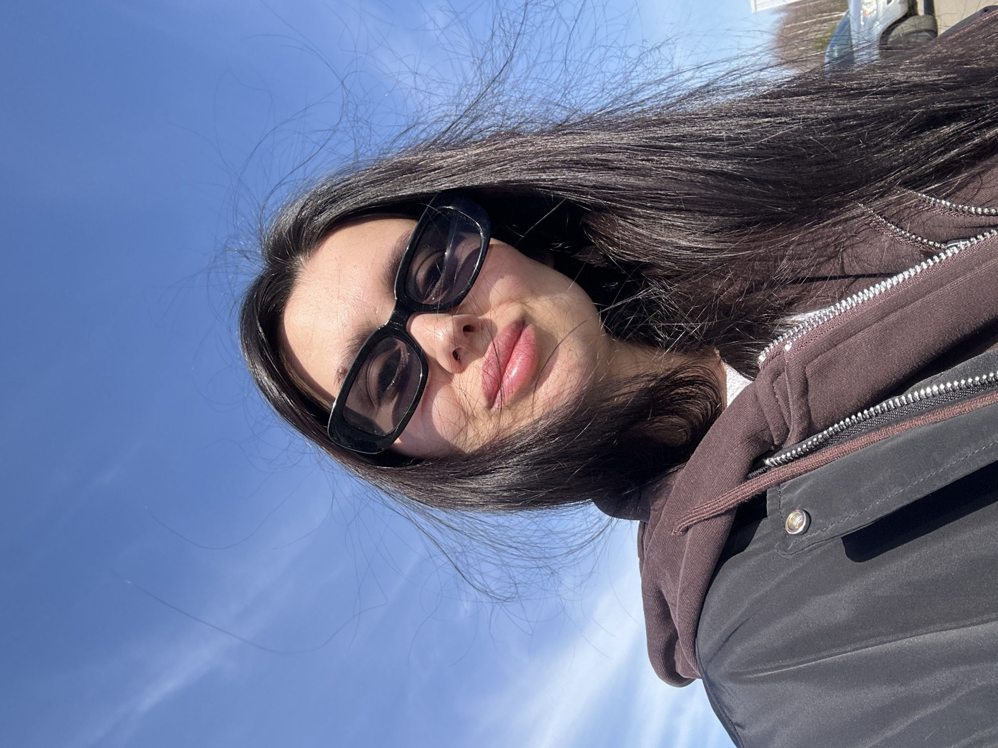 A woman with long dark hair and sunglasses is posing for a selfie outdoors. She is wearing a brown hoodie and a black jacket. The background shows a blue sky with some faint clouds. The location is not discernible as Votkinsk, Russia, or any other specific place based on the provided image.