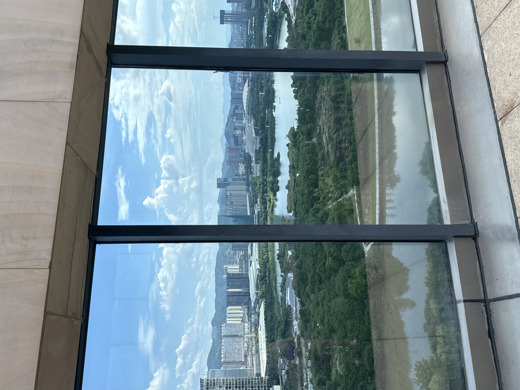 This is an eye-level, medium-shot photograph taken from inside a building looking out through a large, multi-pane window at a cityscape and natural landscape. The window frame is dark grey metal and is divided into three vertical panes. The sky is bright blue with scattered white, fluffy clouds.
In the midground, there is a body of water that reflects the sky and surrounding greenery. A wide, paved pathway runs along the edge of the water. Beyond the water, dense green trees dominate the landscape.
In the background, a city skyline is visible. Tall buildings of various architectural styles, including modern high-rises and some with a more traditional aesthetic, are clustered together. Some buildings are white or light-colored, while others are darker. In the far distance, a range of hazy blue mountains can be seen. There is also a construction crane visible towards the right side of the city skyline.
The foreground of the image shows the interior of the building, with a textured, light-colored ceiling and a window sill made of grey metal and grey tiled flooring.
The scene appears to be in Yiwu, China, likely a park or green space adjacent to an urban development, as indicated by the presence of both natural elements like the lake and trees, and the developing urban infrastructure. There are no people visible in the image. The lighting suggests it is daytime, possibly late morning or early afternoon.
