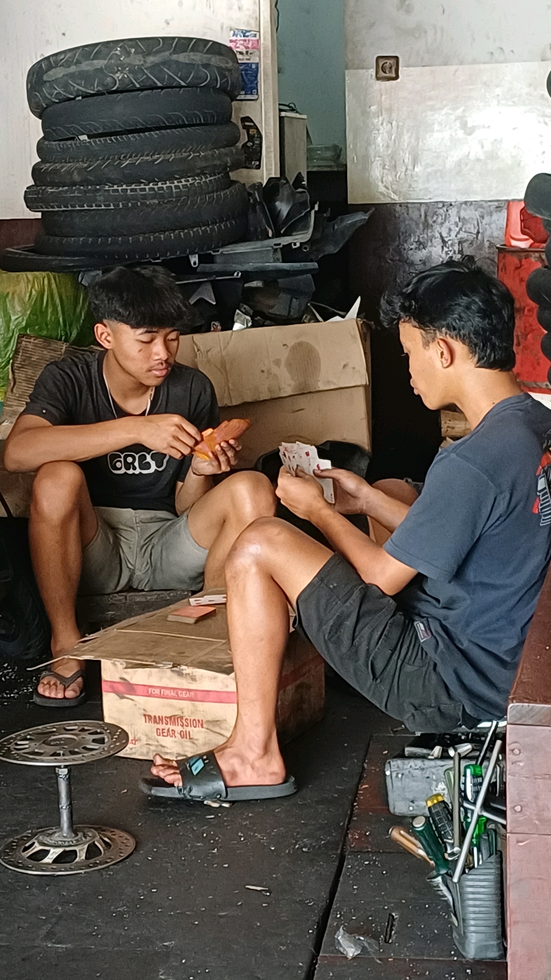Two young men are sitting on the floor of what appears to be a motorcycle repair shop in Temanggung, Indonesia, playing cards. The man on the left is wearing a black t-shirt with "GREY" printed on it and light grey shorts. He is holding a few cards in his left hand and has a small orange object in his right hand. The man on the right is wearing a dark blue t-shirt and black shorts. He is holding a fan of cards in his right hand and is looking at his cards.
The environment is cluttered with various items. To the left, a tall stack of motorcycle tires is visible. In the background, there are cardboard boxes, some tools, and what looks like motorcycle parts. The floor is concrete and dusty. A box labeled "TRANSMISSION GEAR OIL" is visible in the foreground. On the floor to the left, a metallic object resembling a brake rotor on a stand is present. A container of various tools, including screwdrivers and wrenches, is also visible in the lower right portion of the image. The lighting suggests it is daytime, possibly indoors with artificial light.