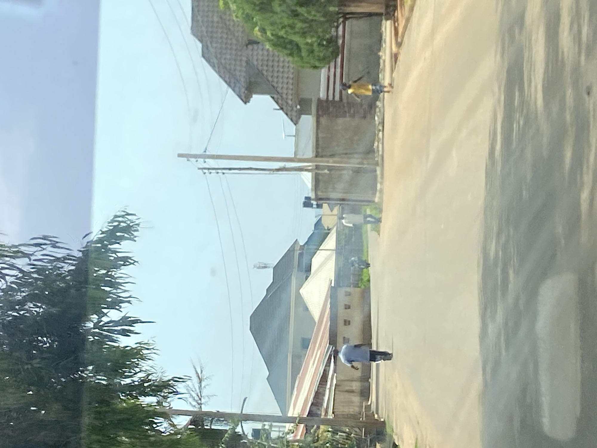 This image captures a street scene in the city of Jos, Nigeria, under a bright, overcast sky. The foreground shows a paved road with tire tracks and scattered shadows from overhead foliage. On the left side of the road, a person wearing a blue shirt and dark pants walks away from the camera. Further down the street, on the right side, a child in a yellow shirt and blue shorts runs towards the right edge of the frame. The background features residential buildings with distinctive rooflines, some appearing modern with pitched roofs and others with flatter structures. Utility poles with electrical wires run along the street. Vegetation, including trees with drooping leaves and a trimmed evergreen tree, frames parts of the scene. The overall impression is a snapshot of daily life on a street in Jos.