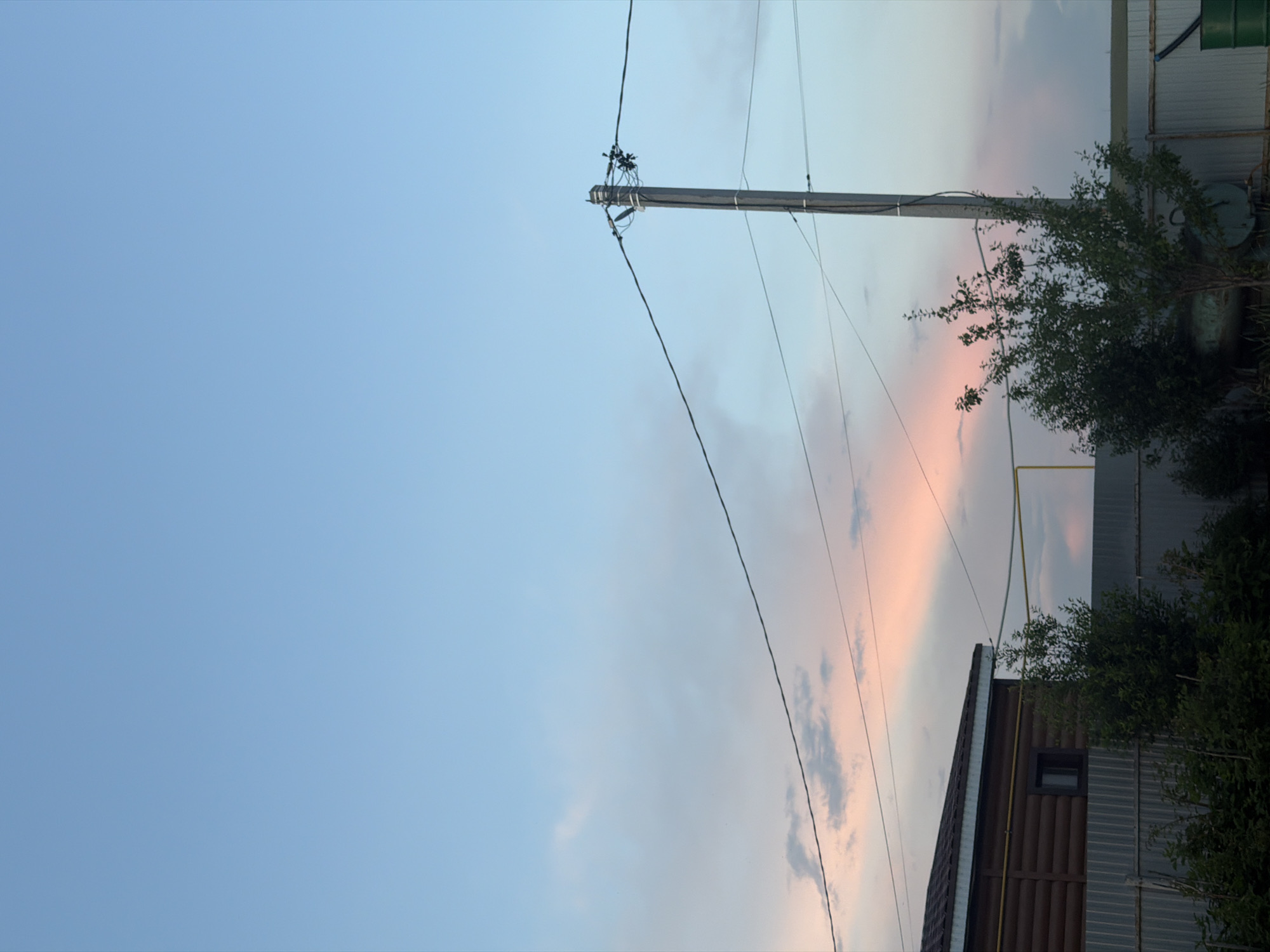 A photograph captures a suburban scene in Nizhny Suyk-Su, Russia, under a pastel-colored sky at dusk. The sky is a gradient of light blue at the top, transitioning to soft pink and pale orange hues near the horizon, with wispy clouds scattered throughout.
In the foreground, a section of a wooden house with a dark brown, horizontally-paneled exterior is visible on the left. It has a dark-framed window and a white trim along the roofline. A grey metal corrugated fence runs horizontally across the lower part of the image, separating the property.
To the right of the house, a wooden utility pole stands tall, supporting multiple black electrical wires that stretch across the frame. A small, leafy green tree is situated in front of the pole and to the right of the fence. Further to the right, a large green barrel is partially visible at the bottom edge of the frame. The overall impression is one of a quiet, rural or semi-rural evening setting.