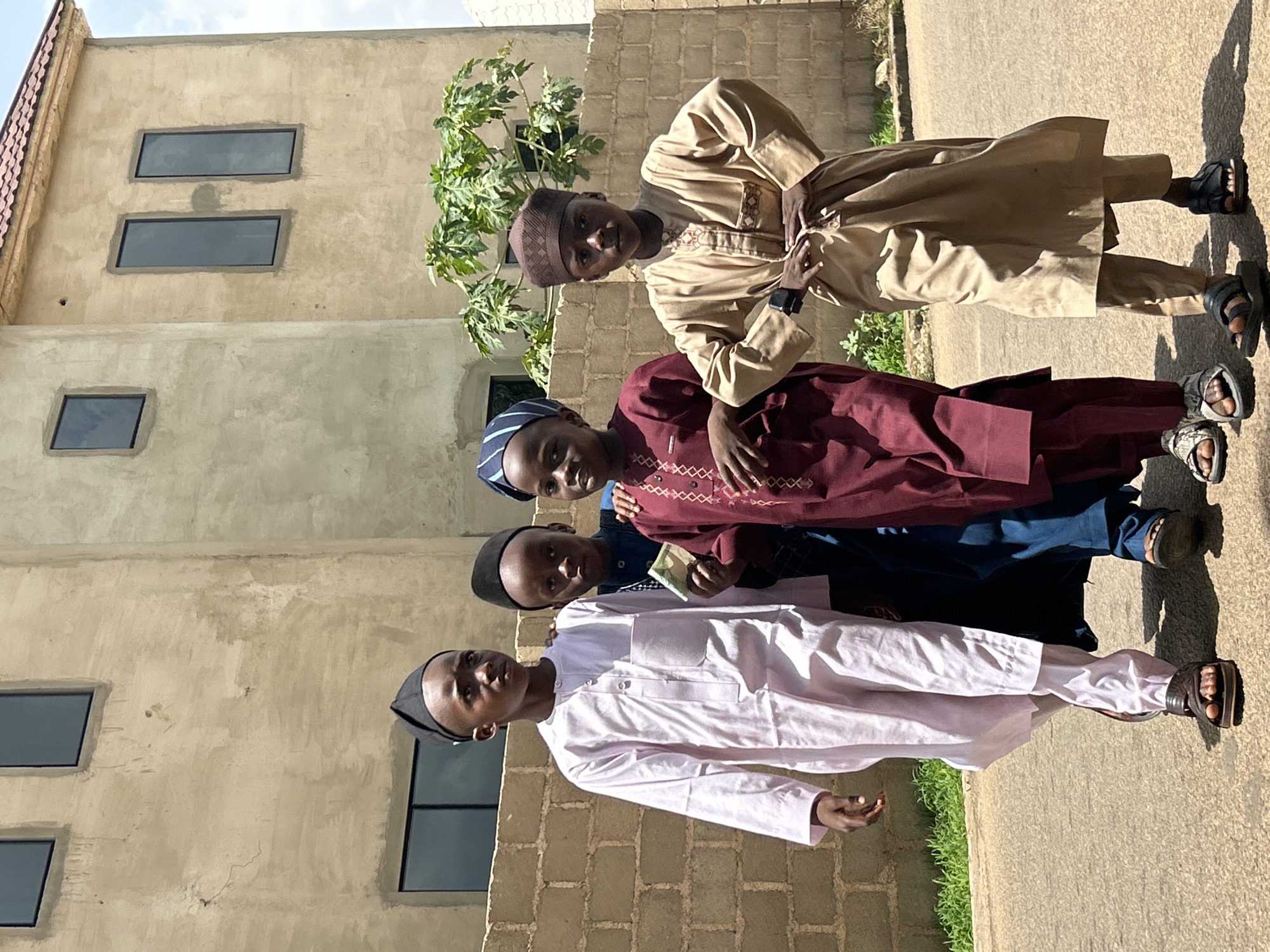 In Jos, Nigeria, four boys are standing on a paved path next to a building. They are dressed in traditional Muslim clothing: the top boy wears a beige robe; the second, a maroon robe; the third, a navy robe; and the fourth, a white robe. The building has a facade of beige brick and stucco, with rectangular windows. A tree is growing next to the building.