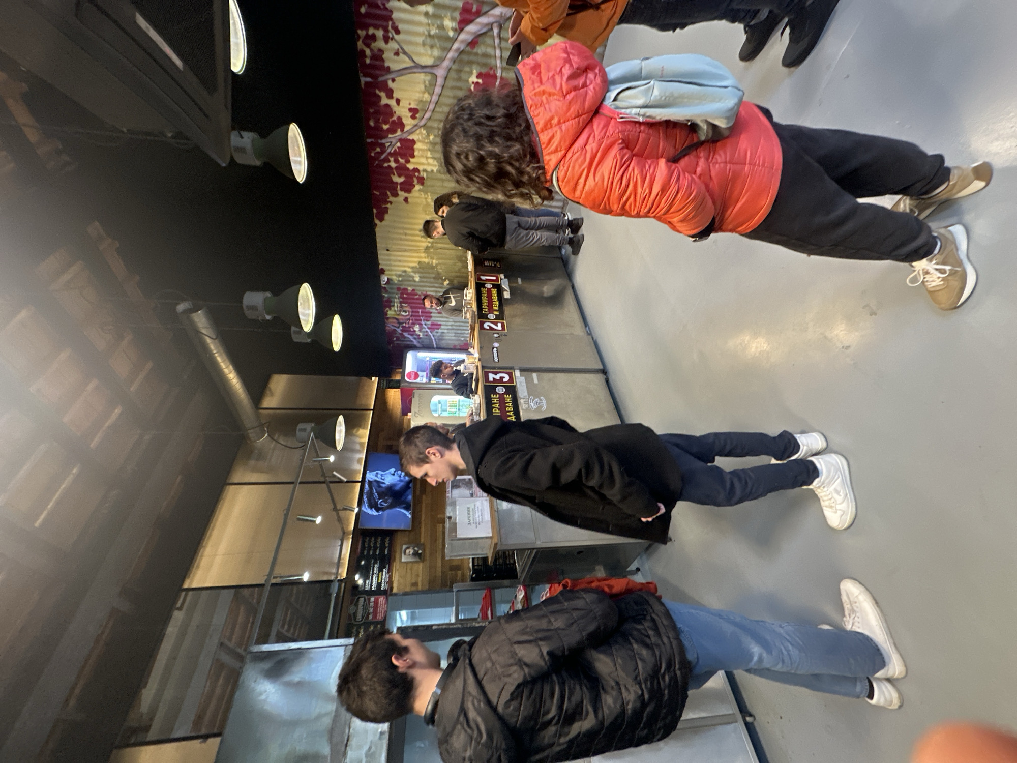 Inside a restaurant located in Stara Zagora, Bulgaria, several people are standing in line at the service counter. A person wearing a red jacket and a light blue backpack is at the front of the line. Behind this individual, a man in a black coat is standing. Further back, another man in a black puffer jacket and jeans is also in line. The service counter has numbered stations, with signage in Bulgarian. The interior of the restaurant features modern lighting fixtures and decorative wall designs.