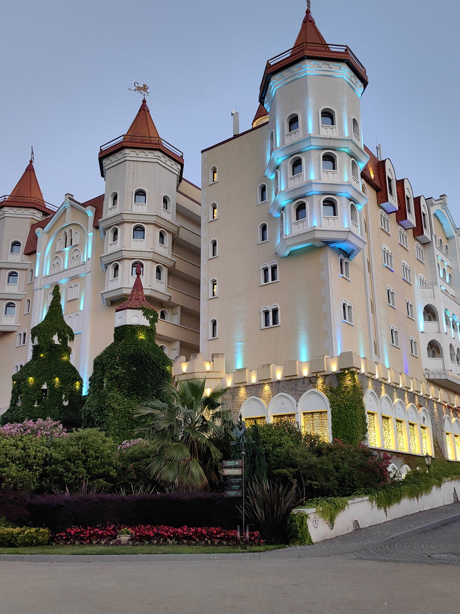 The image shows a large, fairytale-like building with multiple towers and red-tiled roofs, resembling a castle. The building is adorned with blue and yellow lights, suggesting it is illuminated for the evening. The architecture features arched windows and decorative elements. In the foreground, there is a lush garden with various plants, including manicured evergreen topiary shapes, flowering bushes, and palm trees. The garden is bordered by flower beds. The building and its surroundings are well-maintained. The sky is a pale twilight blue, indicating the photograph was taken around dusk. The specific location is identified as being in the city of Sirius, Russia. There are no people visible in the image. The overall impression is one of grandeur and enchantment.