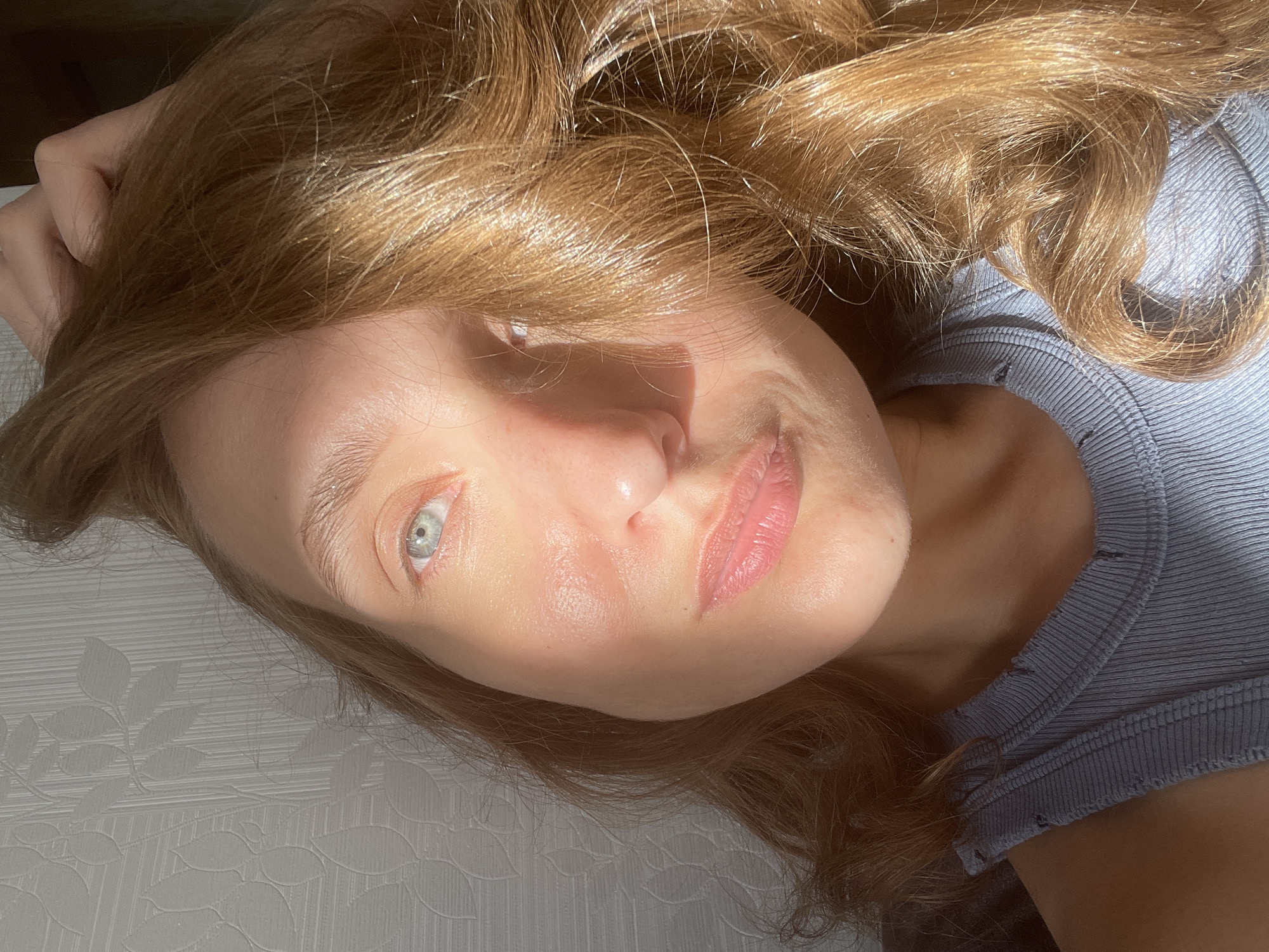 A close-up, rotated portrait features a person, likely a woman, lying down and looking directly at the camera. The subject has fair skin, striking light blue-green eyes, and lustrous golden-brown, wavy hair that frames her face and spreads around her head. Her lips are naturally pink and slightly parted.
Strong, direct natural light, presumably sunlight, illuminates the left side of her face (viewer's right), creating bright highlights on her skin and hair, particularly catching individual strands. The right side of her face is cast in shadow, producing a high-contrast and dynamic lighting effect.
She is wearing a light blue or grey ribbed top, visible on the right side of the image (viewer's left). The background behind her head and to her left is a light-colored, possibly white or off-white, textured fabric with a subtle leaf or floral pattern, suggesting an indoor setting such as a bed or couch. No text is visible within the image. The photograph conveys a calm and direct gaze, captured in bright, natural light. This media file is associated with the location context of Sharkan, Russia.