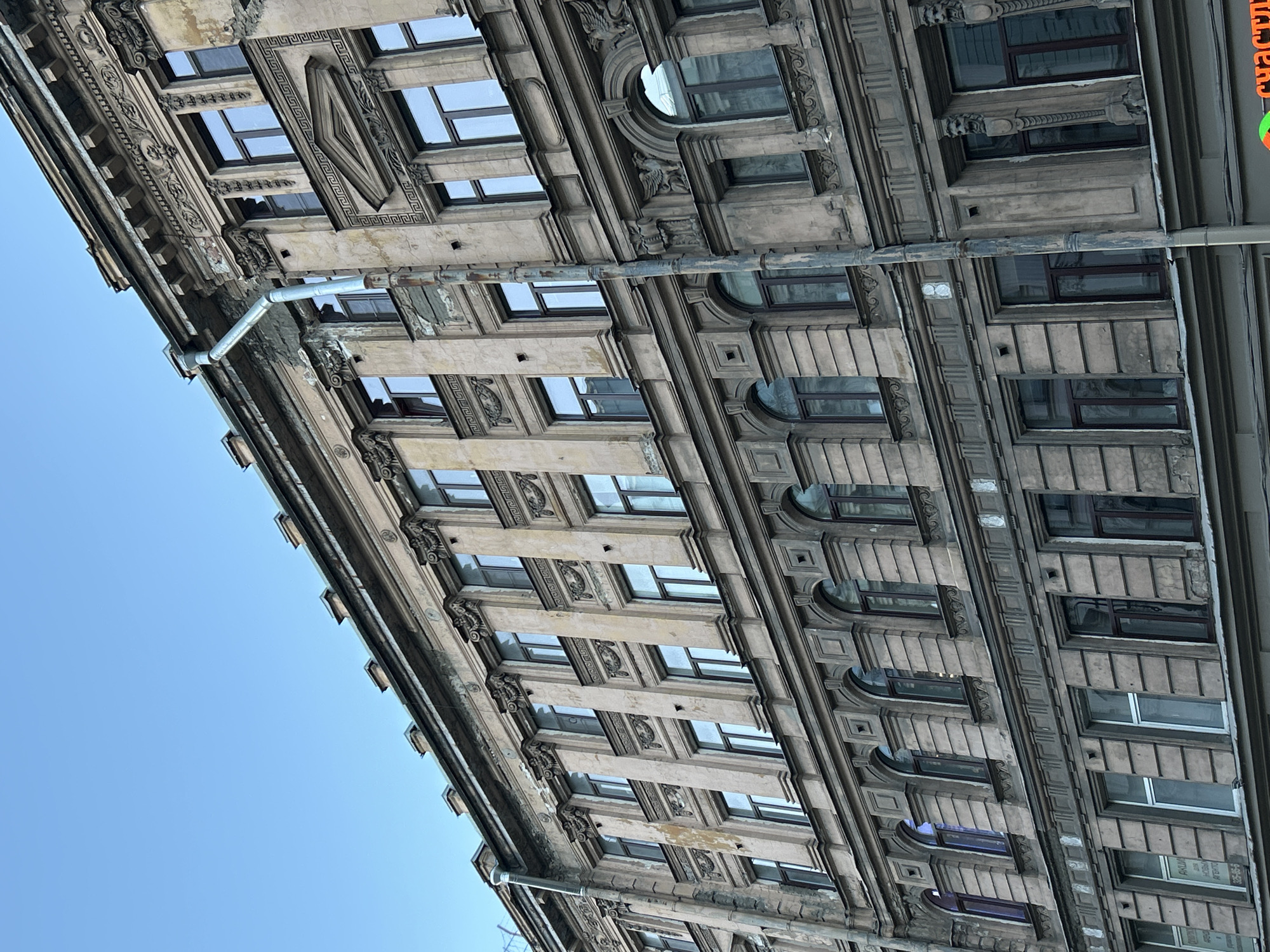 The image captures a detailed, upward-angled view of an ornate, multi-story building facade against a clear blue sky, likely taken from street level. The building, consistent with the architectural style found in Saint Petersburg, Russia, displays classical or neoclassical elements.
The facade is rich with intricate details, including decorative stone carvings, moldings, pilasters, cornices, and friezes featuring Greek key patterns and stylized faces or masks. Windows vary in shape, with many being rectangular, some having arched tops, and at least one circular window visible on a higher floor. The building appears aged and weathered, with visible discoloration, flaking plaster, and general wear on its stone or stucco surfaces.
A prominent, rust-stained metal downspout or pipe runs vertically along the facade, collecting water from a gutter system at the roofline. The clear, bright blue sky in the upper-left portion of the frame indicates daytime and fair weather conditions.
In the lower-right section of the building, on a window, some Cyrillic text is visible. Partial words can be discerned, such as "КУРИА" (KURIA) and what appears to be a phone number or partial address "295-5-" or "5-55-". Another small sign has the word "счастли" (schastlivi, meaning happy/fortunate). No people or explicit activity are depicted, the focus is solely on the architecture.
