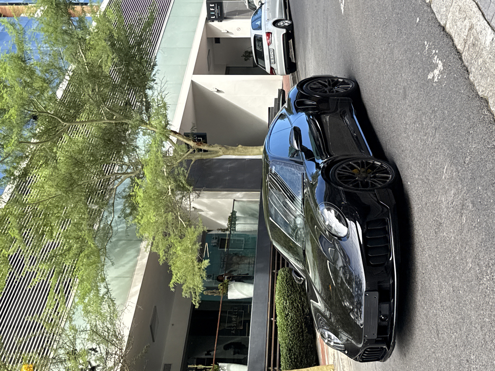 A black Porsche 911 parks on a grey paved street in front of a modern building in Cape Town, South Africa. A tree with green foliage is visible on the left side of the frame. A white Audi is parked further down the street. The building has a combination of glass and architectural grill elements. "LIFE" is visible on the facade. In the windows of the building is the name ASSIRELLI.