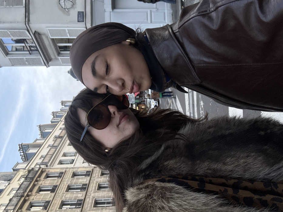 Two women pose close together on a city street. One woman, wearing sunglasses and a fur-trimmed coat, looks toward the camera. The other woman, in a headscarf and brown leather jacket, puckers her lips. Tall buildings line the street, and people walk in the background.