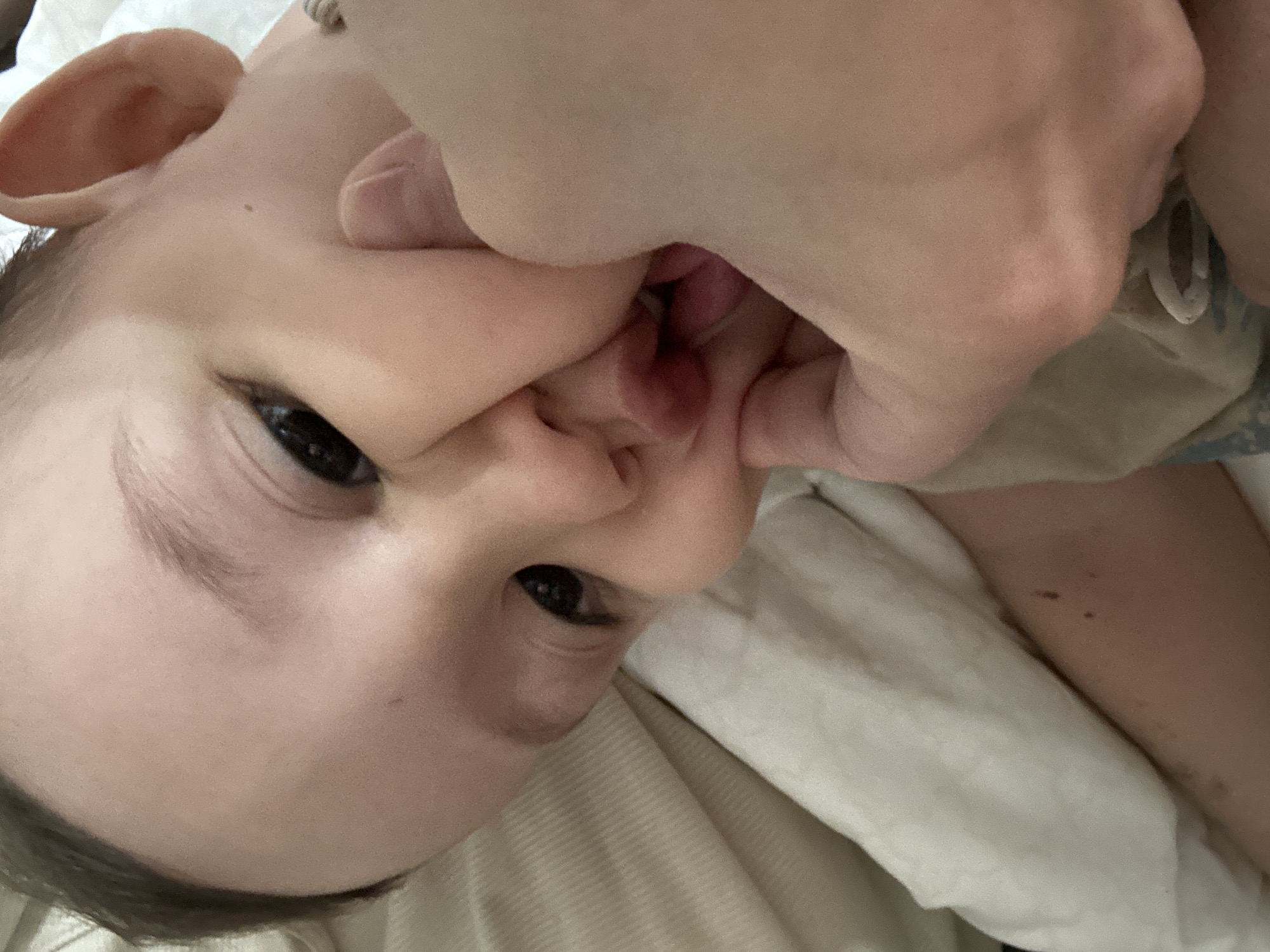 A close-up, rotated image shows a baby with its face being manipulated by an adult hand. The baby has fair skin, dark hair, and dark eyes. An adult hand is positioned over the baby's mouth, seemingly pulling at its cheeks. The background consists of a light-colored fabric, possibly bedding. The image provides no information relating to Chaikovsky, Russia.