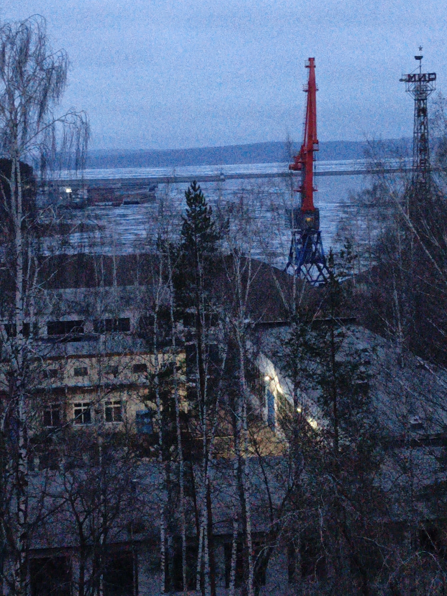 This photograph captures a stark, low-light scene in Chaikovsky, Russia, likely at dusk or dawn during winter or late autumn. The image has a grainy texture, contributing to its somewhat somber mood.
In the foreground, a dense array of bare deciduous trees, including slender birches with their pale trunks and branches, and some darker conifers, obscure parts of the view. Their leafless state suggests cold weather.
Beyond the trees, architectural elements emerge. A multi-story building, possibly residential or industrial, is visible in the mid-left, with several windows emitting a warm, artificial light that contrasts with the cool blue tones of the environment.
The upper midground is dominated by two tall, man-made structures. On the left, a prominent red industrial crane stands tall against the sky, its metallic framework dark blue at the base. To its right, a tall, dark metal lattice tower rises, topped with a distinct star. Clearly visible on this tower are the Cyrillic letters "МИР" (MIR), which translates to "peace" or "world."
In the background, a wide body of water, likely the Kama River or Votkinsk Reservoir, extends across the horizon. The surface of the water appears partially frozen or covered in ice, with darker stretches indicating open water or reflections. The sky above is a uniform, pale blue-grey, suggesting an overcast condition. No human activity is discernible, lending a quiet and still atmosphere to the cold, semi-industrial landscape.
