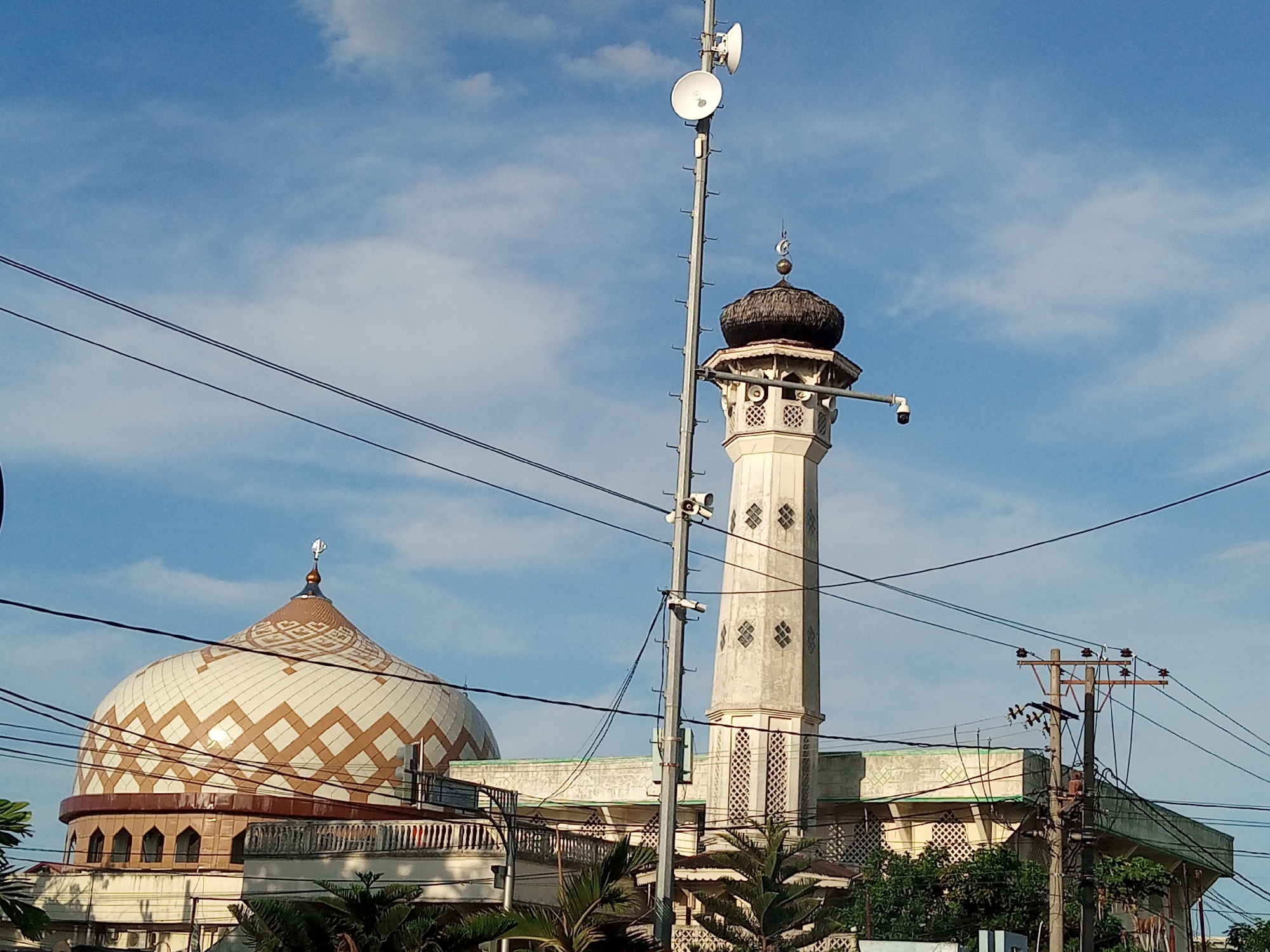 The image depicts a mosque in Lhokseumawe, Indonesia, under a blue sky with scattered clouds. The mosque features a large, patterned dome and a tall minaret with a dark-colored dome and Islamic crescent symbol. A tall, metallic pole equipped with speakers is positioned in front of the minaret. Overhead power lines crisscross the frame. The mosque structure is surrounded by greenery, including trees and smaller plants. An electrical pole is visible on the right side of the frame. The overall impression is of an urban environment with religious architecture integrated into the cityscape. A security camera is mounted near the top of the minaret.
