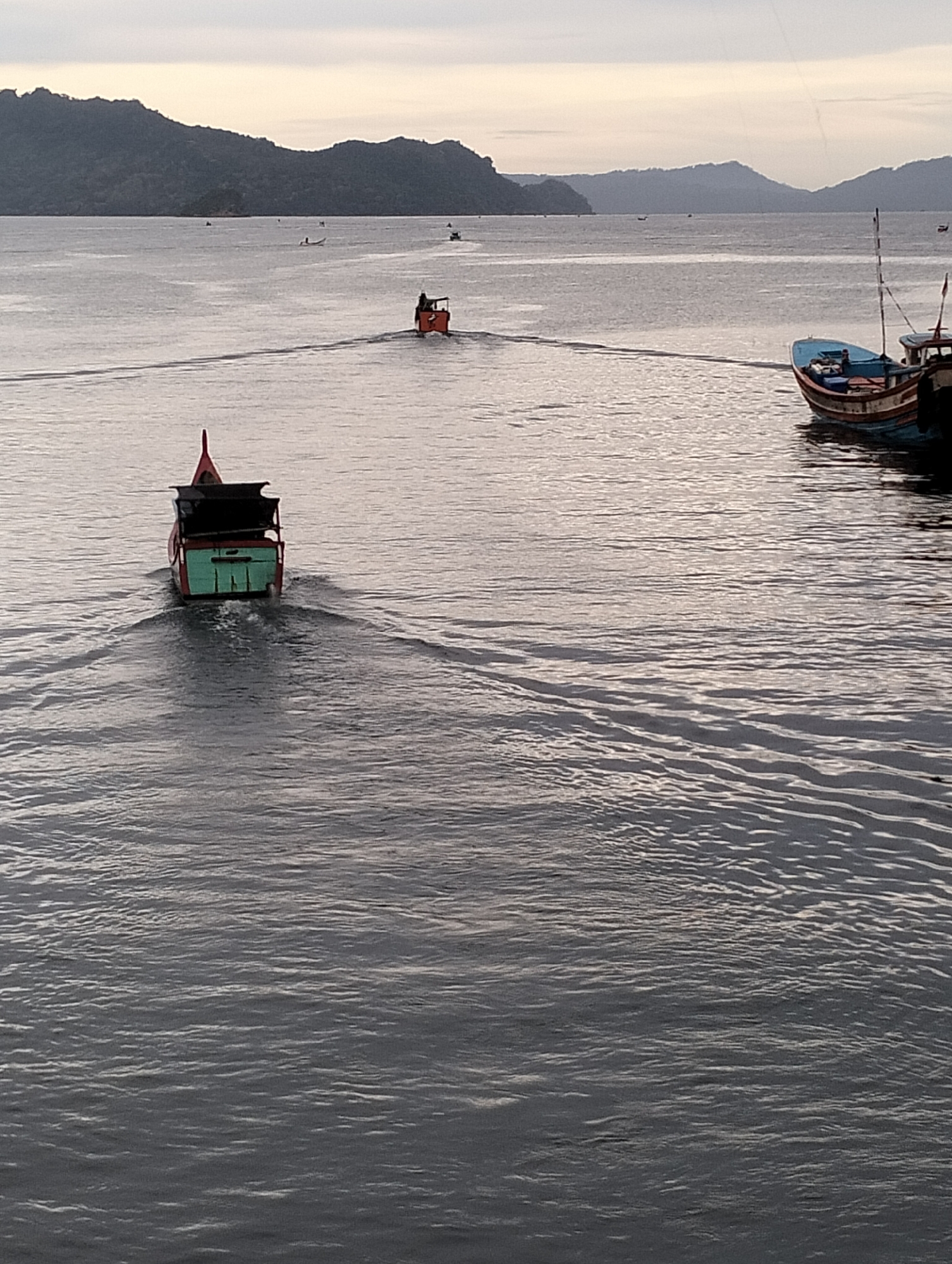 The image depicts a seascape in Banda Aceh, Indonesia, likely near a coastal area or harbor. In the foreground, a small green and red boat is moving away from the viewer, leaving a wake on the water's surface. Further in the distance, another small orange boat is heading straight away from the viewer. To the right side of the image, are two moored boats. The water appears calm with gentle ripples and reflections. In the background, there is a mountainous coastline under a gray sky.