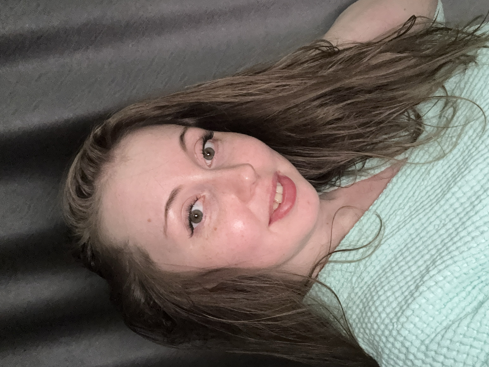 The image captures a young woman lying down in an indoor setting, looking directly at the camera with a relaxed expression. She has long, wavy brown hair, which appears slightly damp or naturally textured, spread around her head and shoulders. Her head is tilted, and she presents a soft, closed-mouth smile, with her lips slightly parted to reveal a hint of her teeth. Her eyes are open and appear to be hazel (brownish-green), with visible eyelashes. Her fair skin shows a few faint freckles across her cheeks.
She is wearing or covered by a light teal or mint green, textured garment, possibly a blanket or a top with a waffle-knit pattern. The background behind her head is a dark grey, textured surface, likely a sofa or bed. Another portion of the light teal textured material is visible on the right side of the frame, blending with her attire. The lighting is soft and even, suggesting a comfortable indoor environment without harsh shadows. The overall mood conveyed is casual and friendly. No text is visible in the image.