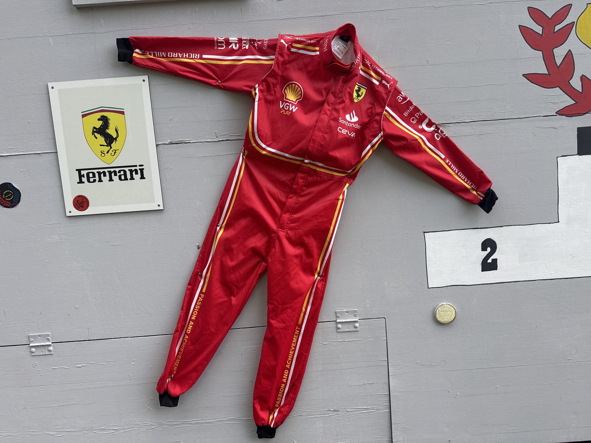This image shows a red Ferrari racing suit hanging on a grey wooden wall. The suit is adorned with various logos, including "Richard Mille," "VGW PLAY," "Shell," "Santander," and "CEVA LOGISTICS." The Ferrari Prancing Horse logo is visible on the chest and on a separate sign to the left of the suit. This sign also displays the "SF" logo and the word "Ferrari" in black lettering. To the right of the suit, a white rectangle with the number "2" in black is affixed to the wall. Below this is a small circular metallic object. The overall setting appears to be an outdoor display, possibly a roadside attraction, located in Nendeln, Liechtenstein.