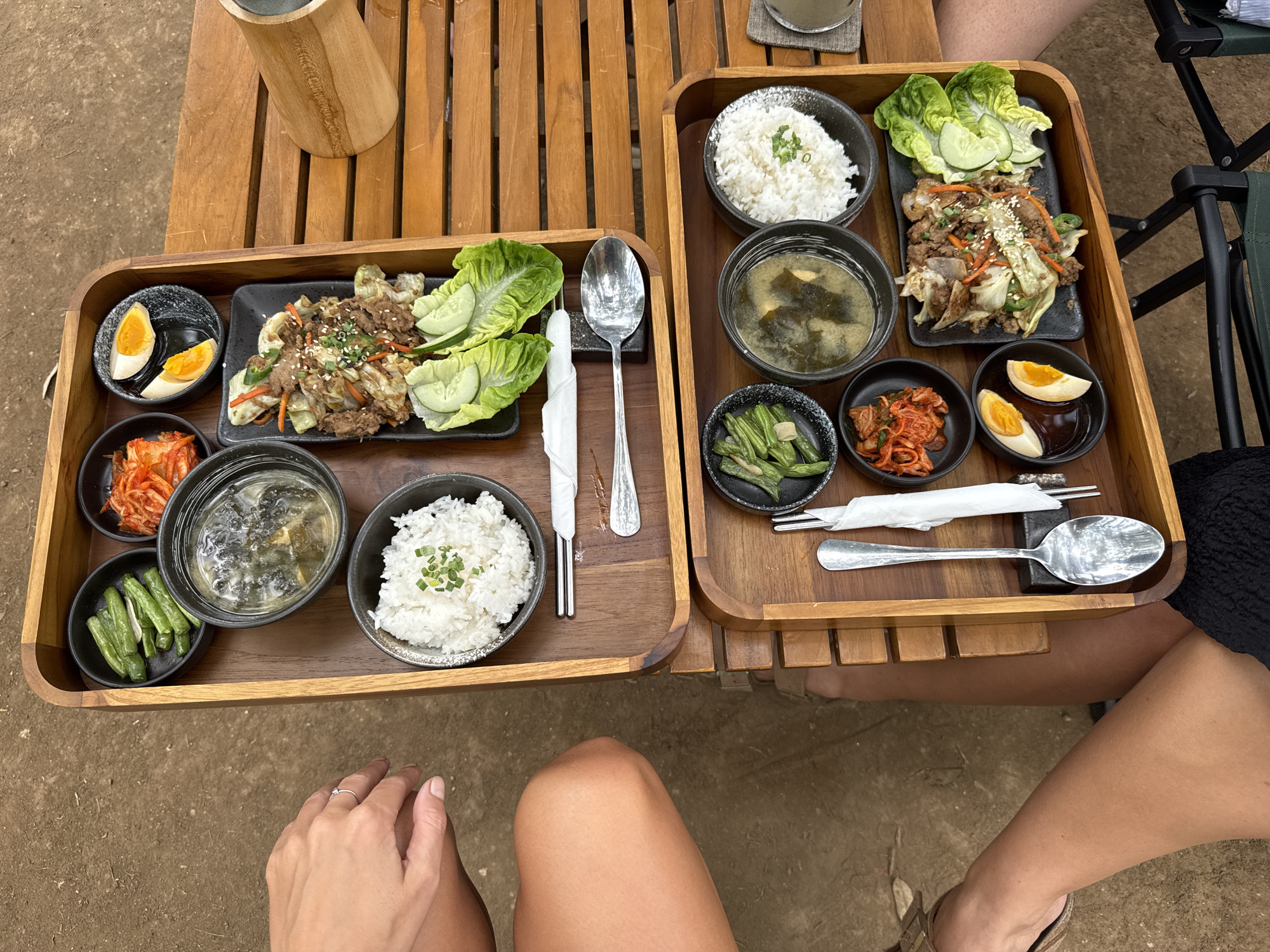 Two wooden trays of Korean food are placed on a slatted wooden table outdoors, suggesting a casual dining setting. The trays contain identical meals, each consisting of a bowl of white rice, a bowl of soup, a small plate of green beans, a small dish of kimchi, a plate of sliced hard-boiled eggs with dipping sauce, and a main dish of bulgogi with lettuce wraps and cucumber slices. Utensils, including chopsticks and spoons, are provided for each meal. The ground around the table appears to be dirt or gravel, typical of an outdoor area. Two individuals are partially visible in the frame; one's hands are resting on the upper tray, and the other's legs are visible on the left and bottom of the frame. The overall scene suggests two people enjoying a meal together in an outdoor restaurant.